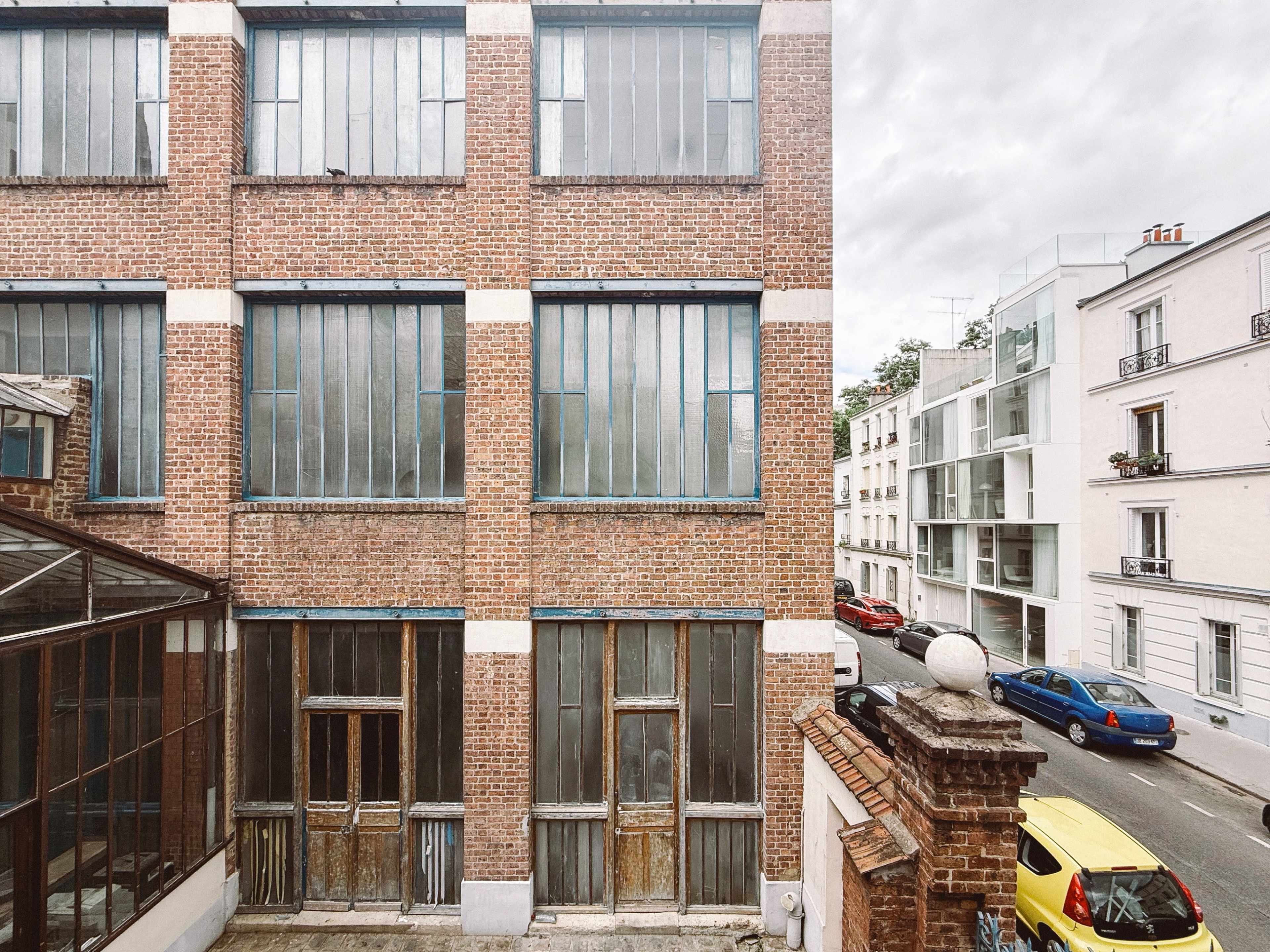 The image shows a brick building with large, barred windows next to a modern white building and parked cars on a street.