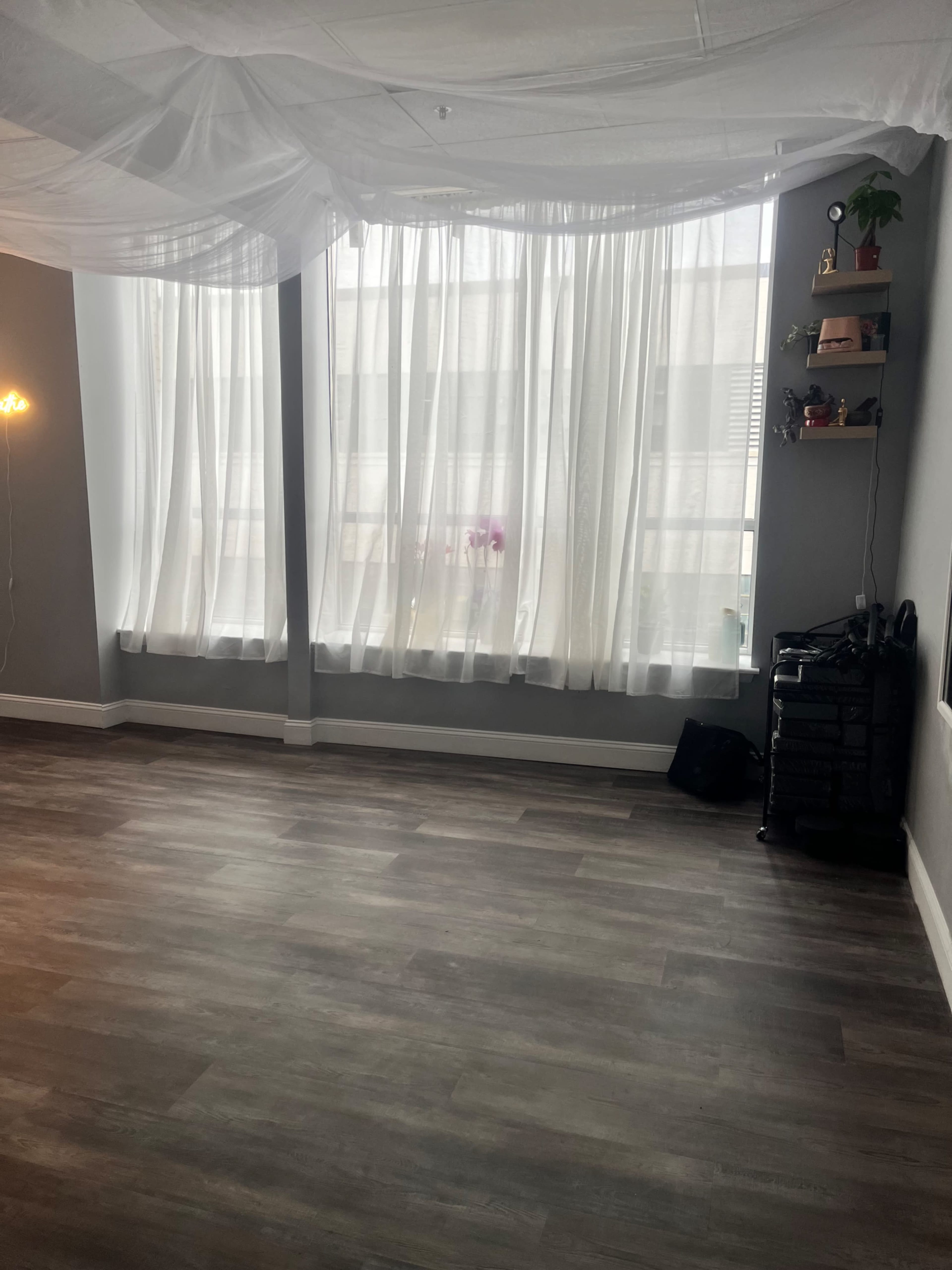 A spacious room features large windows covered with sheer white curtains, allowing soft light to filter through, with light wood flooring and minimal decor.