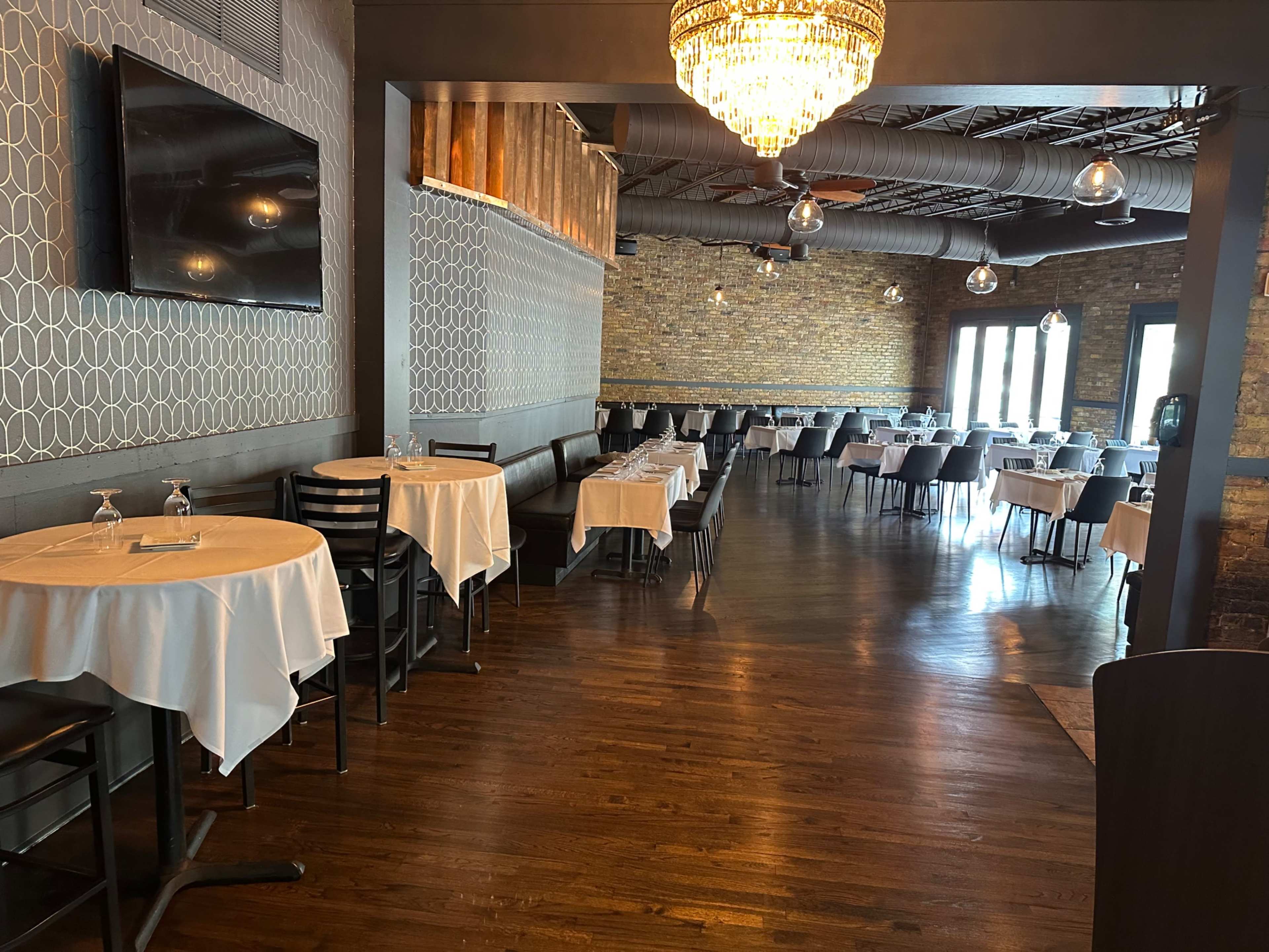 The image shows a spacious restaurant interior featuring neatly set tables with white tablecloths, a brick wall, and a chandelier overhead.
