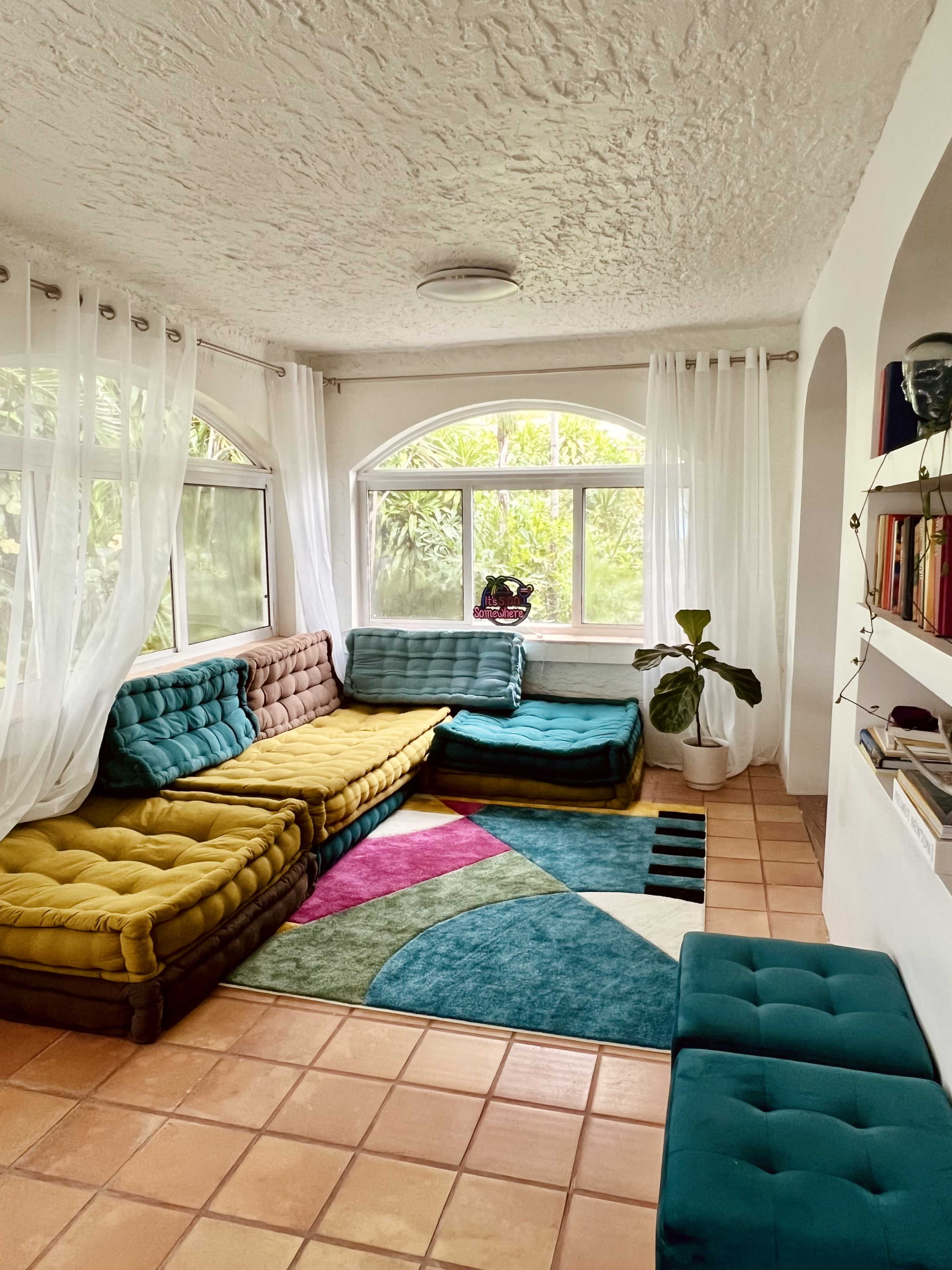 A bright, airy living room with large windows, a colorful sectional sofa, and a rug featuring geometric patterns.