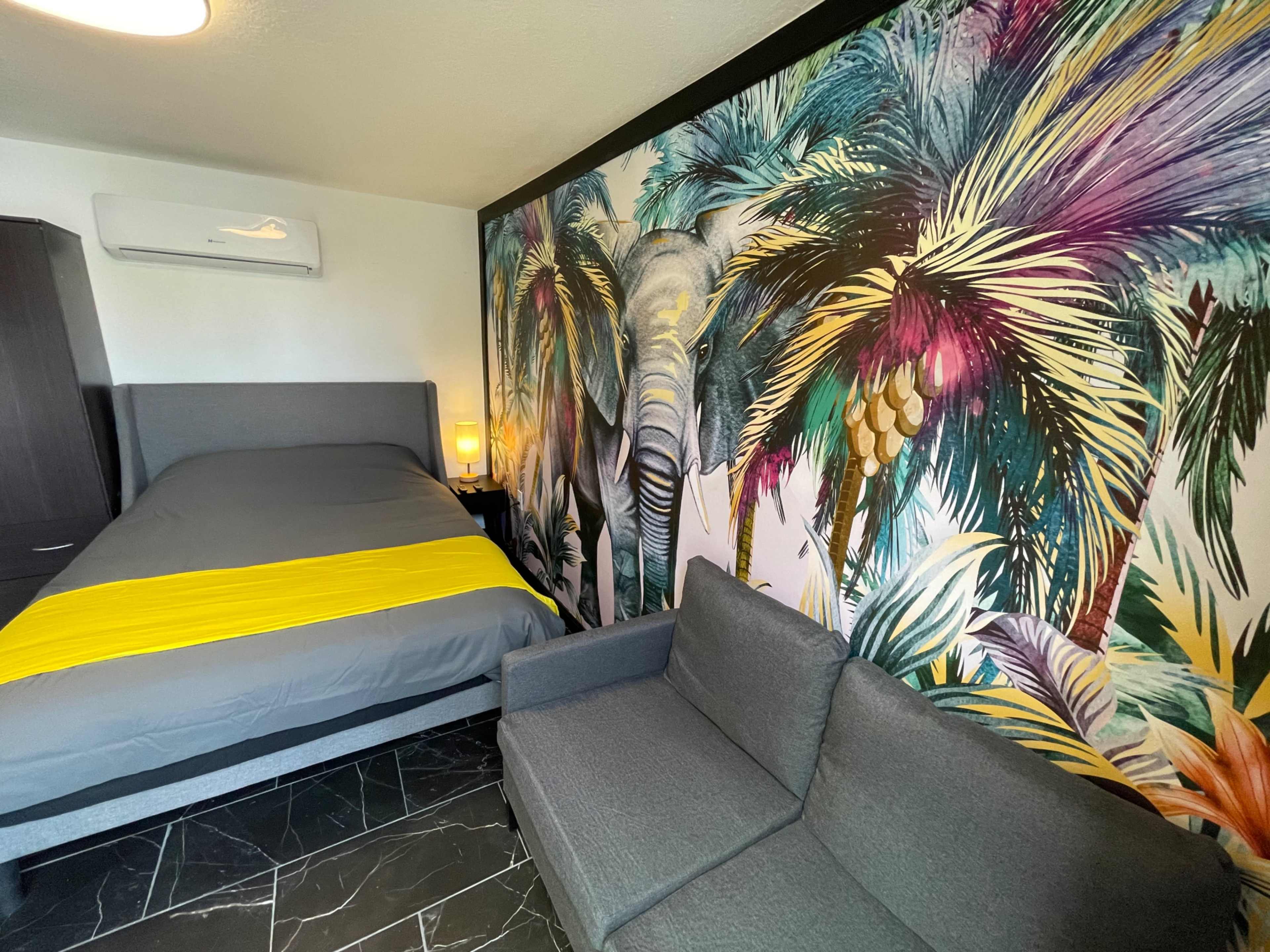 A bedroom featuring a gray bed with a yellow blanket, a couch, and a colorful mural of elephants and palm trees on one wall.