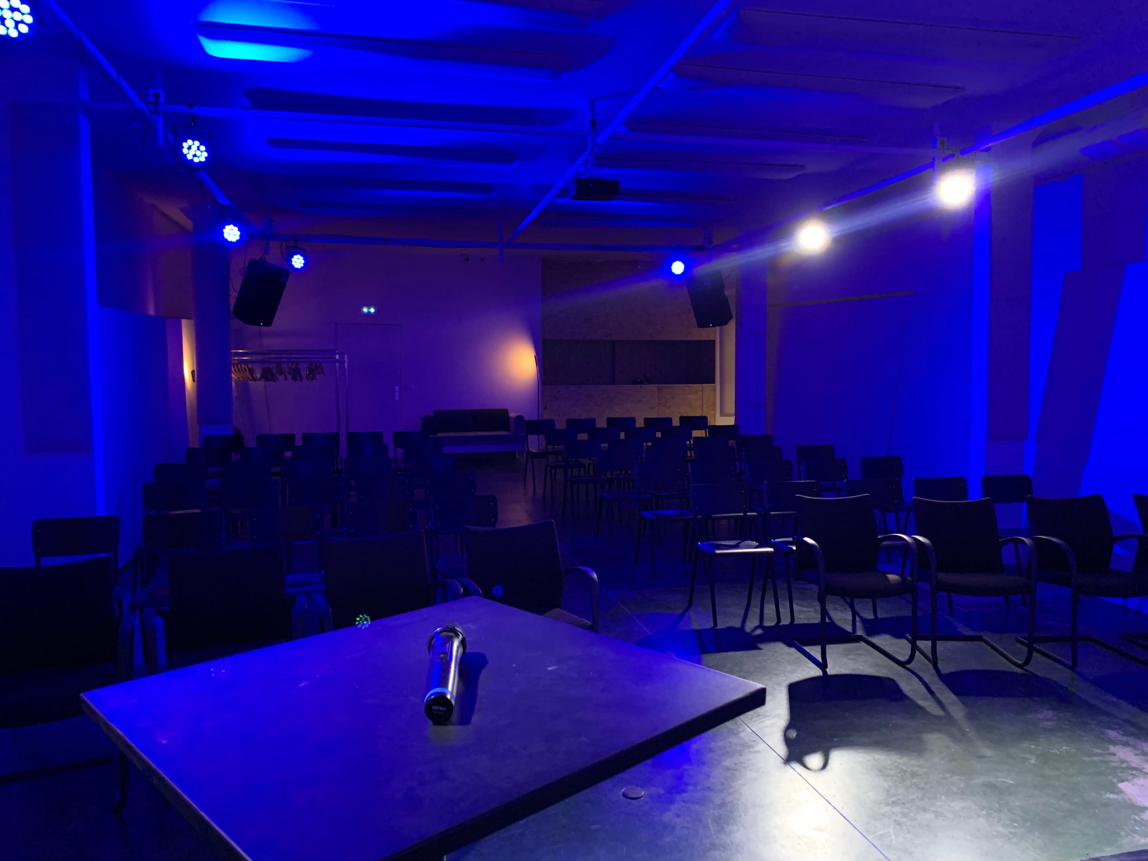 A dimly lit auditorium features rows of black chairs facing a central table under blue stage lighting.