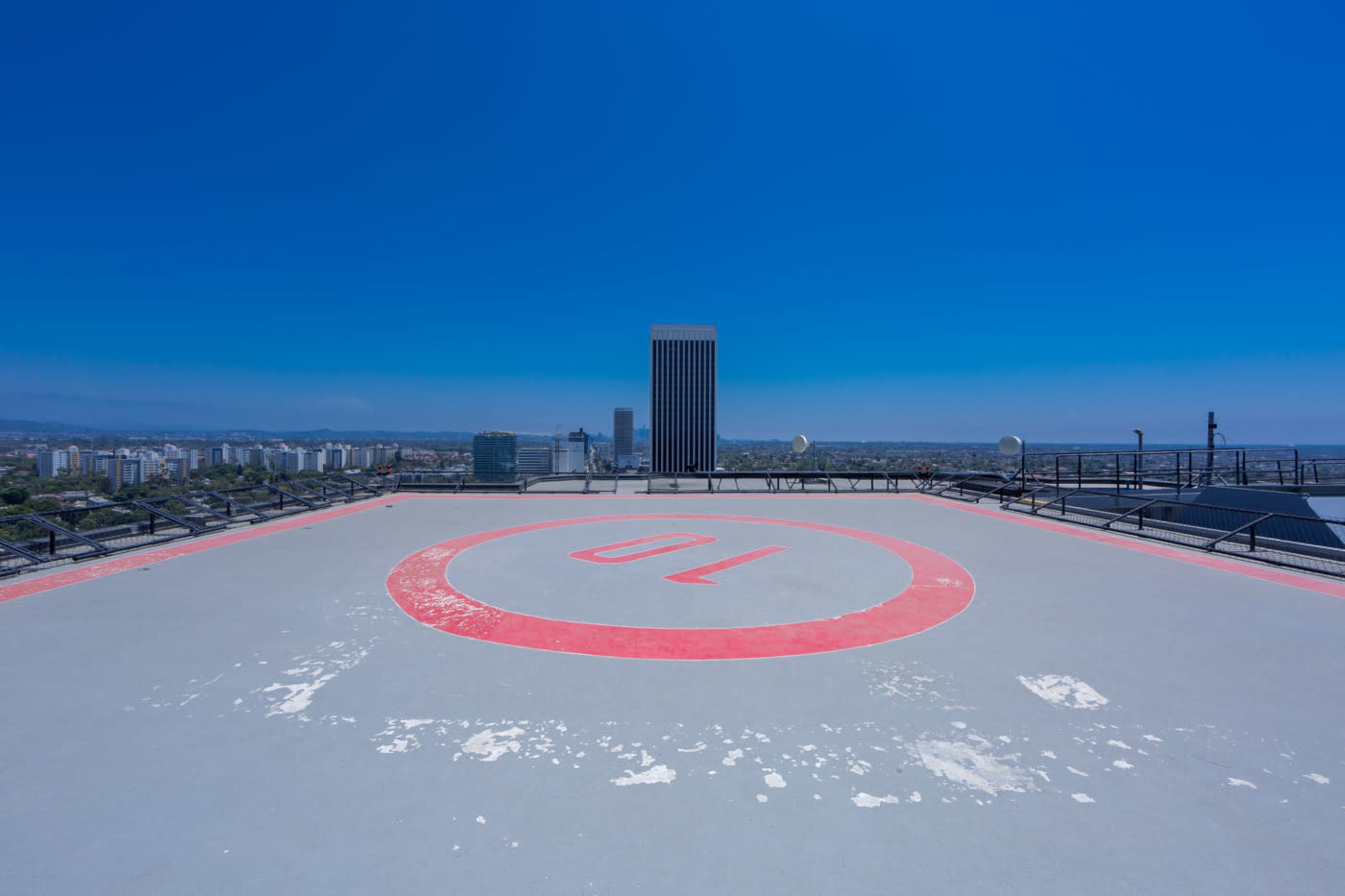 The image shows a helicopter landing pad on a rooftop with a red circle marked "10" and a city skyline in the background under a clear blue sky.