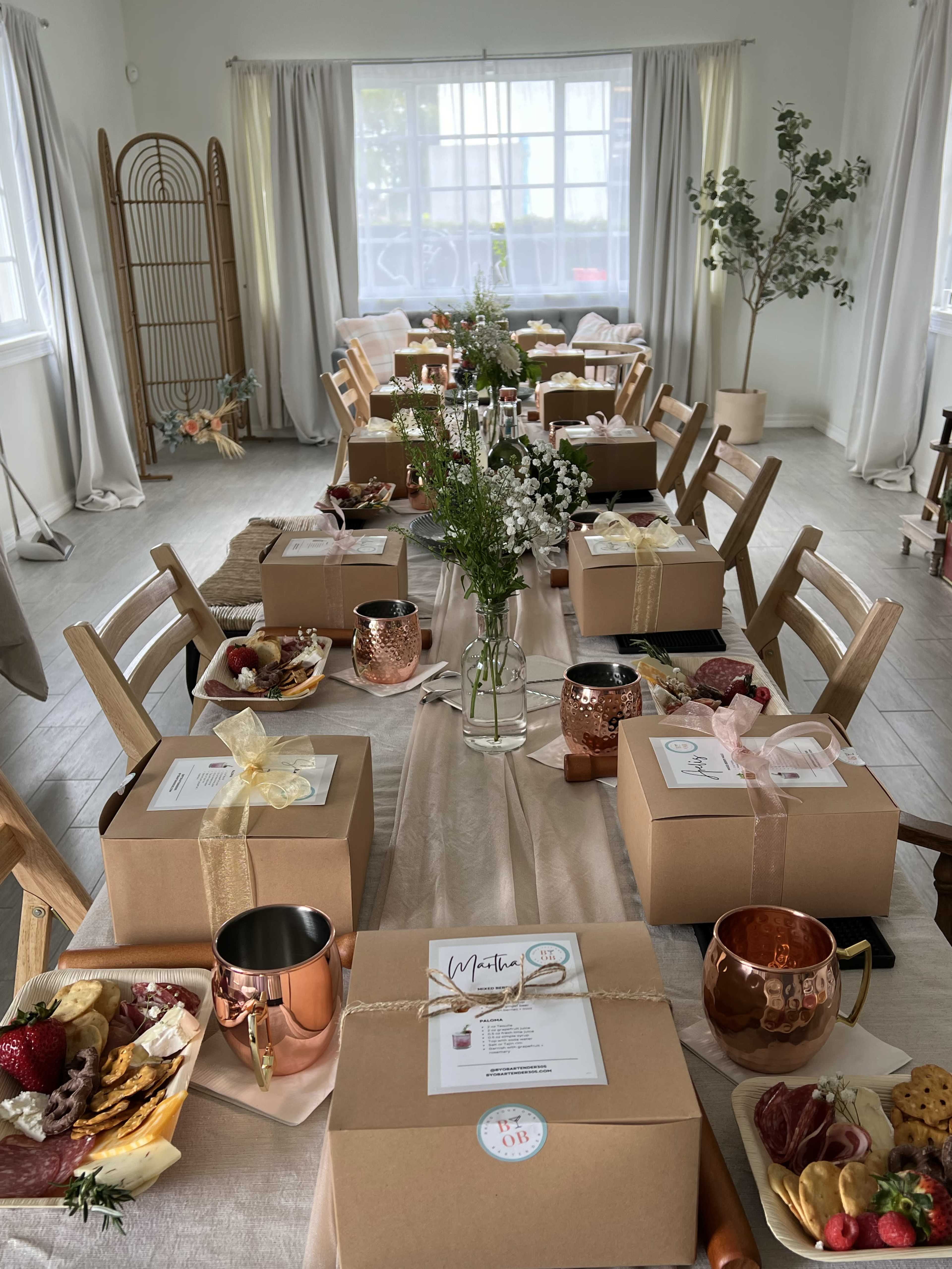 A dining area is set with multiple tables covered in beige tablecloths, featuring neatly arranged gift boxes, decorative flowers, and various snack platters.