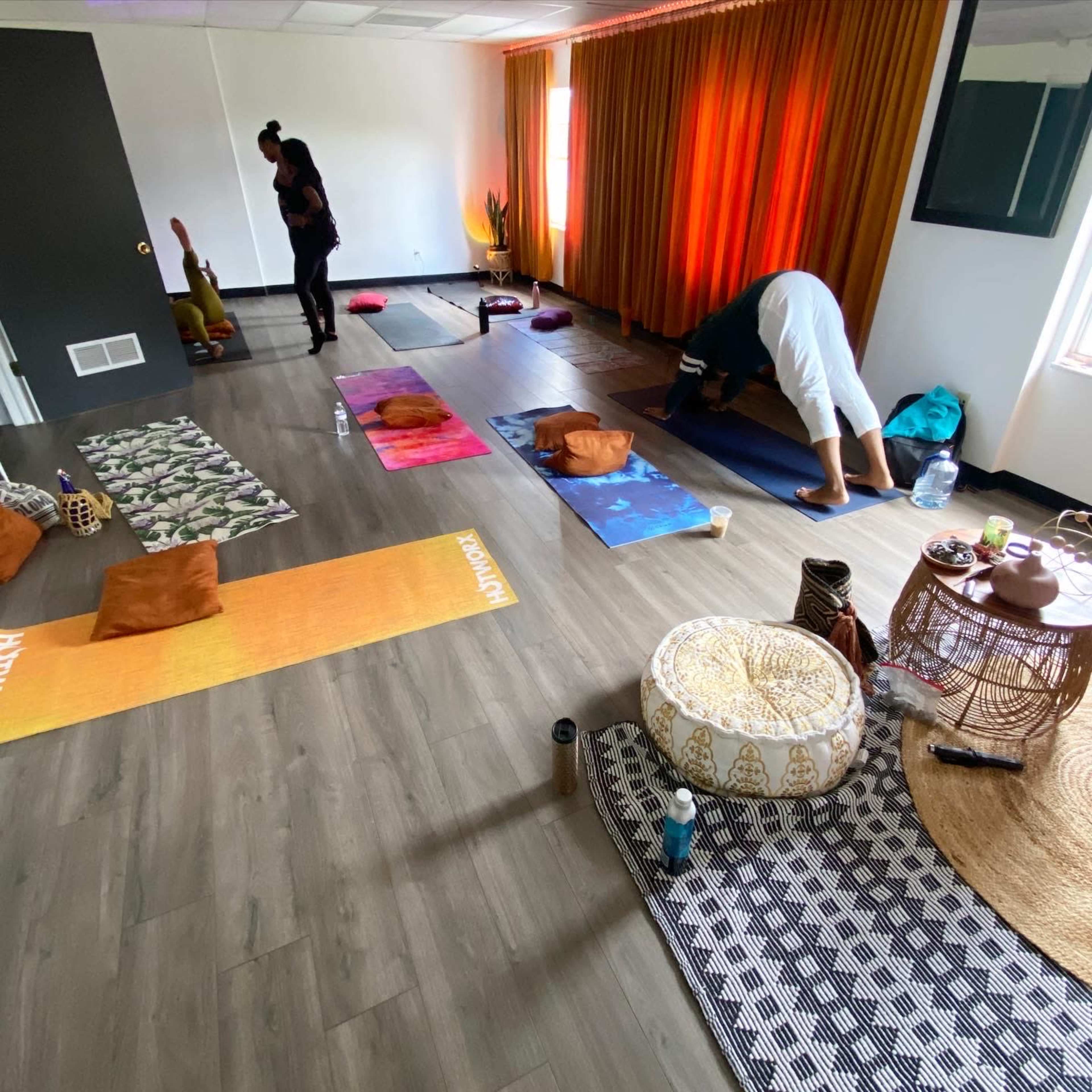 A spacious room is set up for a yoga session, featuring mats, cushions, and a variety of decorative textiles, while two individuals prepare for practice.