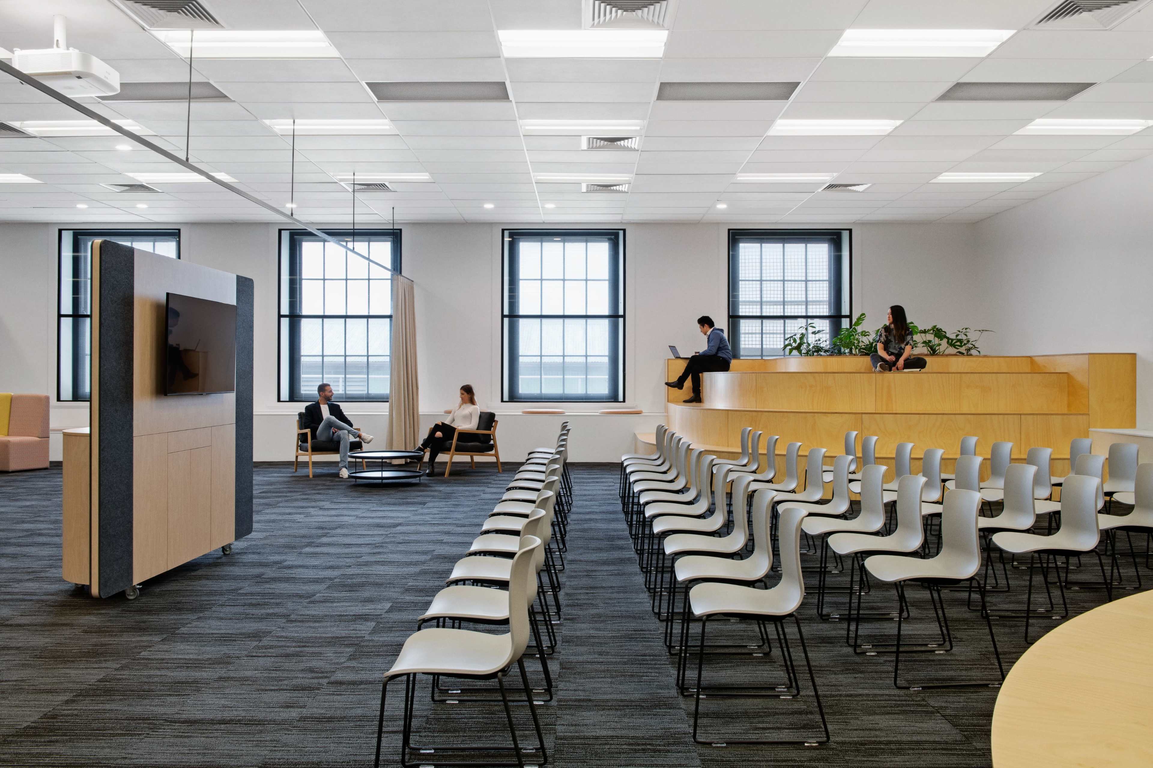 A modern, minimalist office space with rows of chairs facing a large wooden panel that houses a TV, complemented by seating areas and large windows.