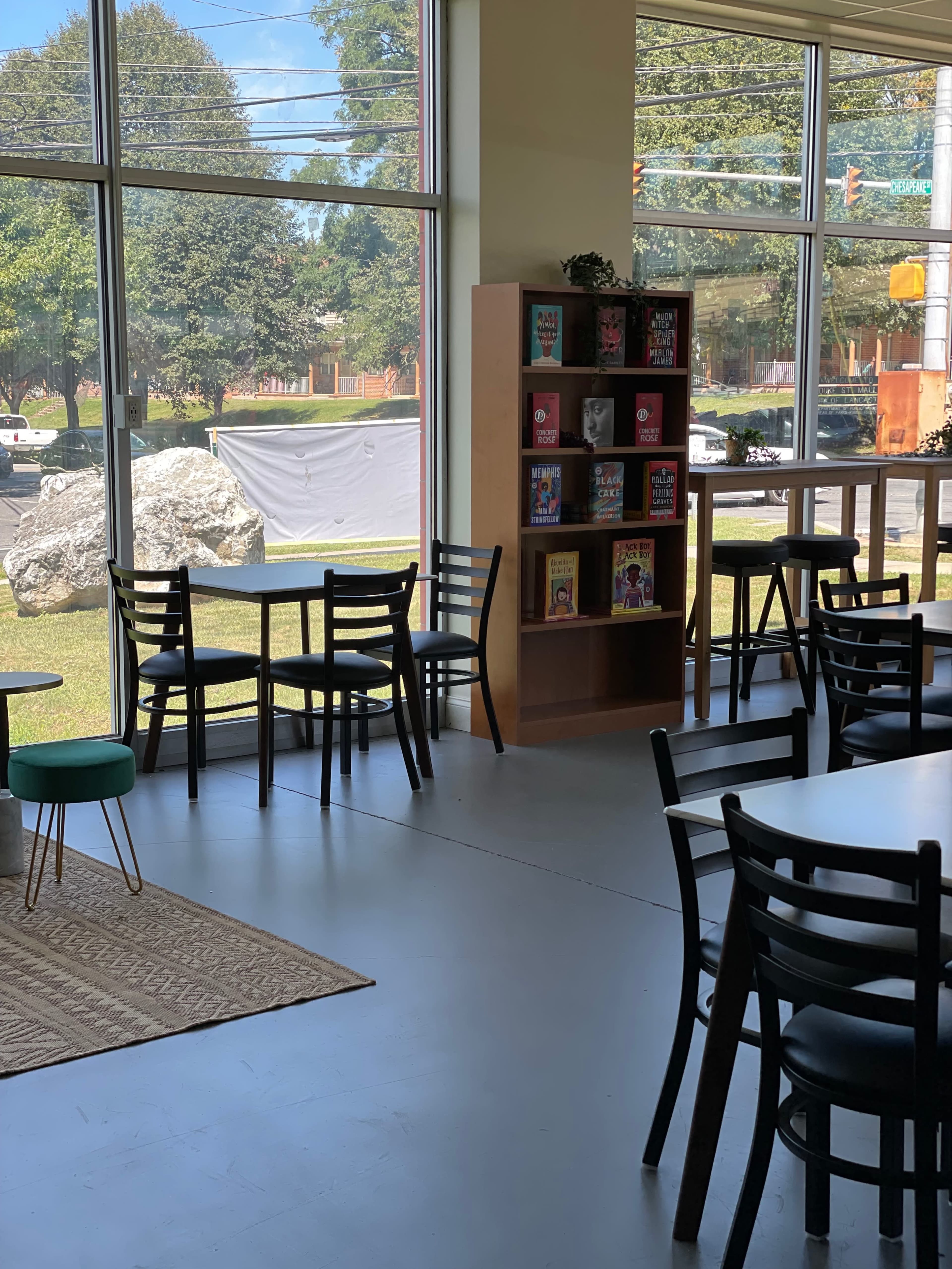 A bright interior space with empty chairs and tables, a bookshelf filled with colorful books, and large windows overlooking a grassy area outside.