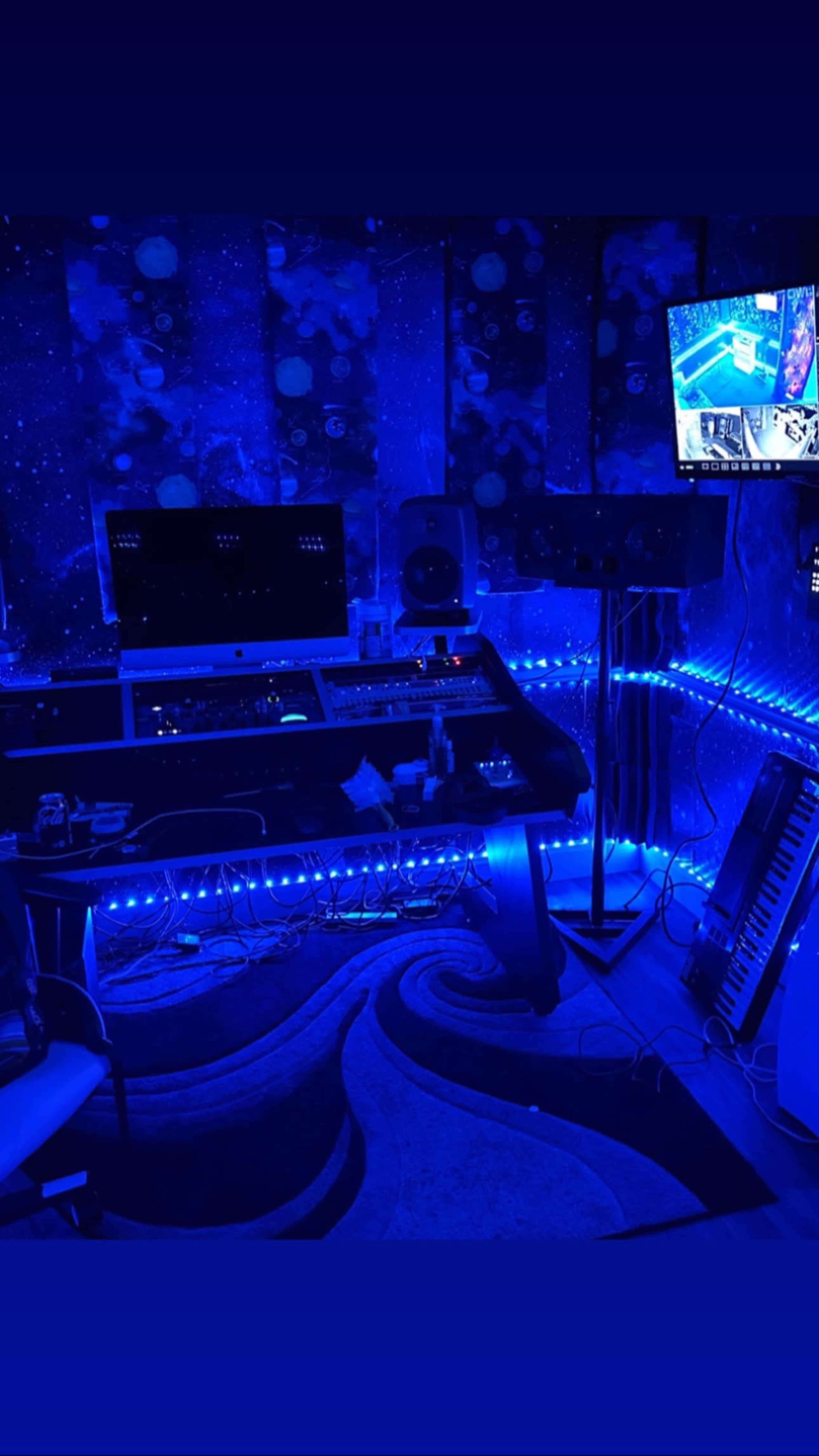 A dimly lit music studio with blue LED lights, an audio mixing desk, a computer, and a television displaying video content.