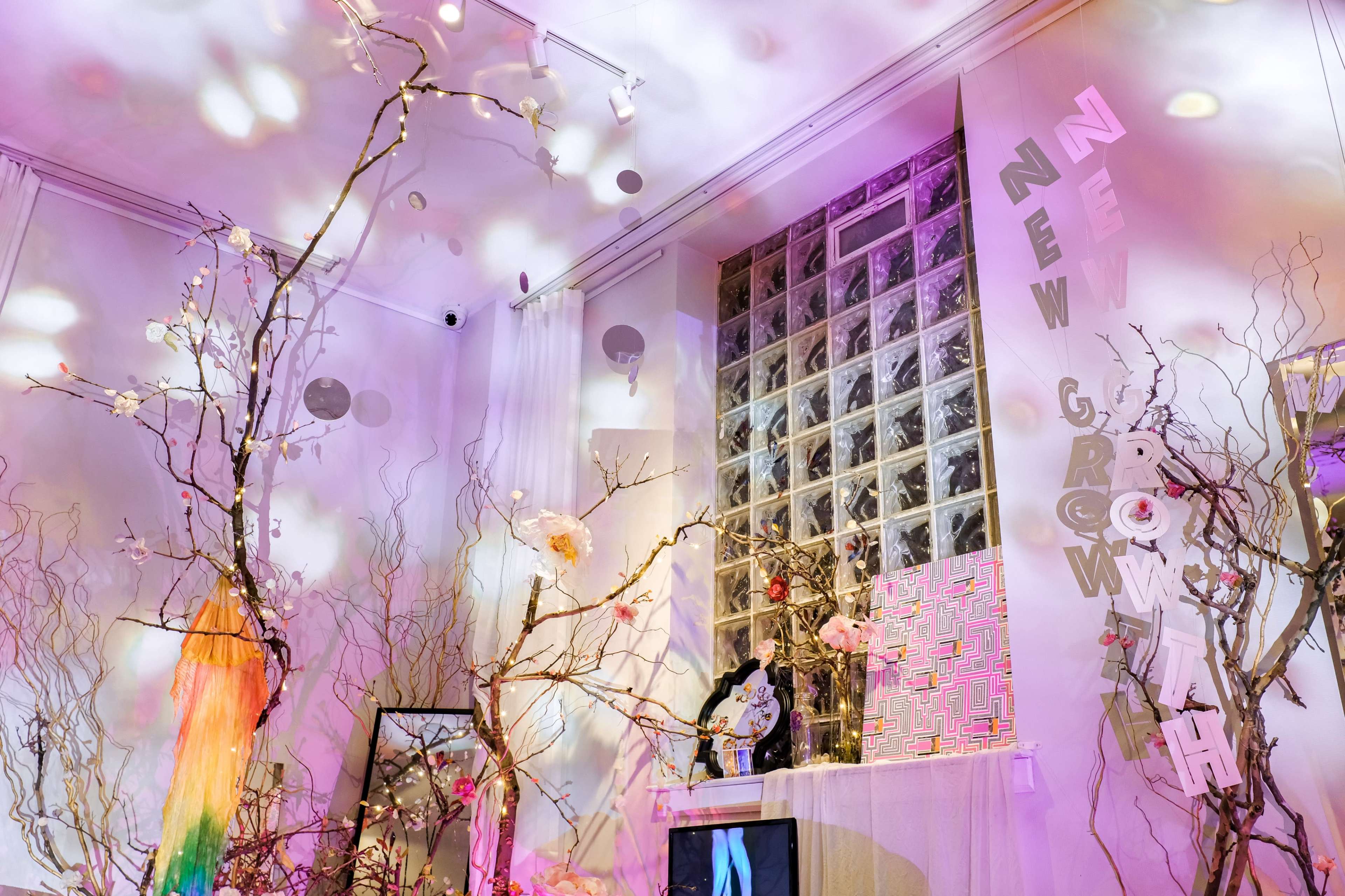 The scene features a room decorated with bare branches and colorful lights, showcasing the words "NEW GROWTH" on the wall and a vibrant artwork on display.