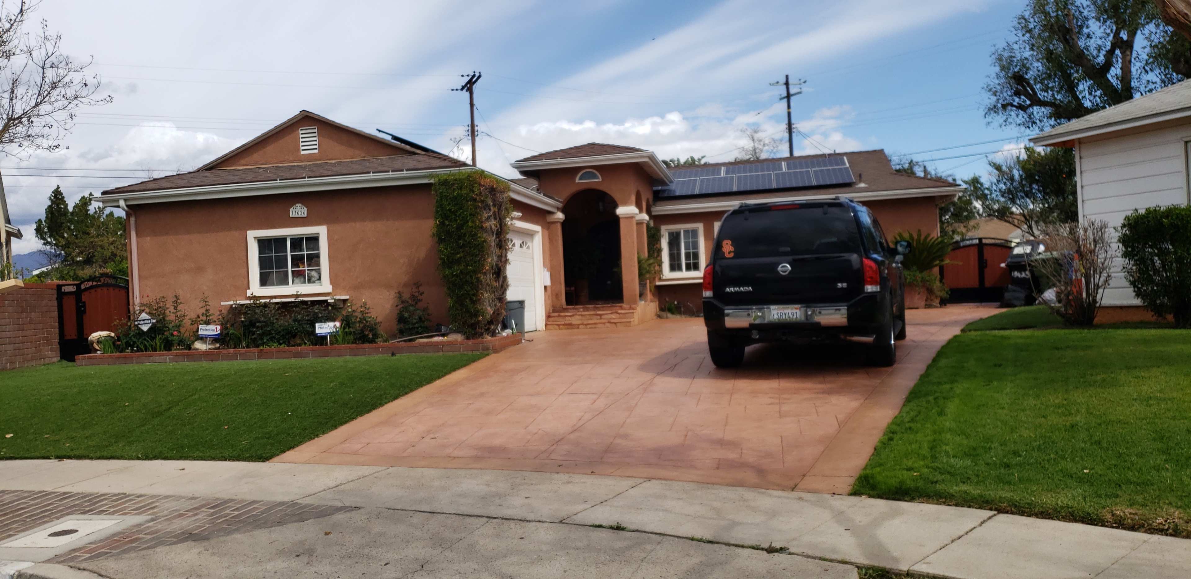 A single-story house with a brown exterior and a front driveway is flanked by a black SUV parked on a paved surface.