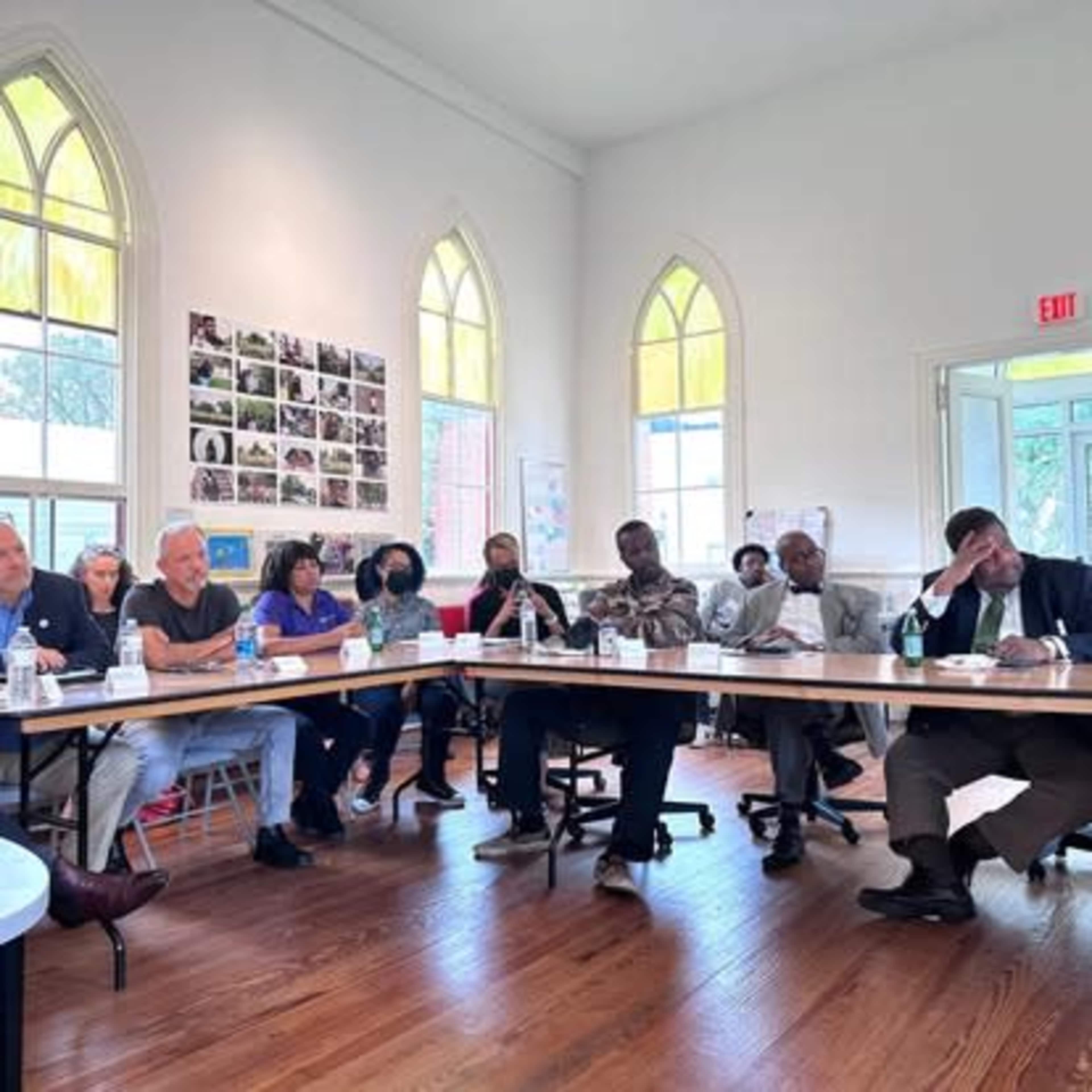 A group of people is sitting around a long table in a brightly lit room, engaged in discussion, with various materials and documents in front of them.