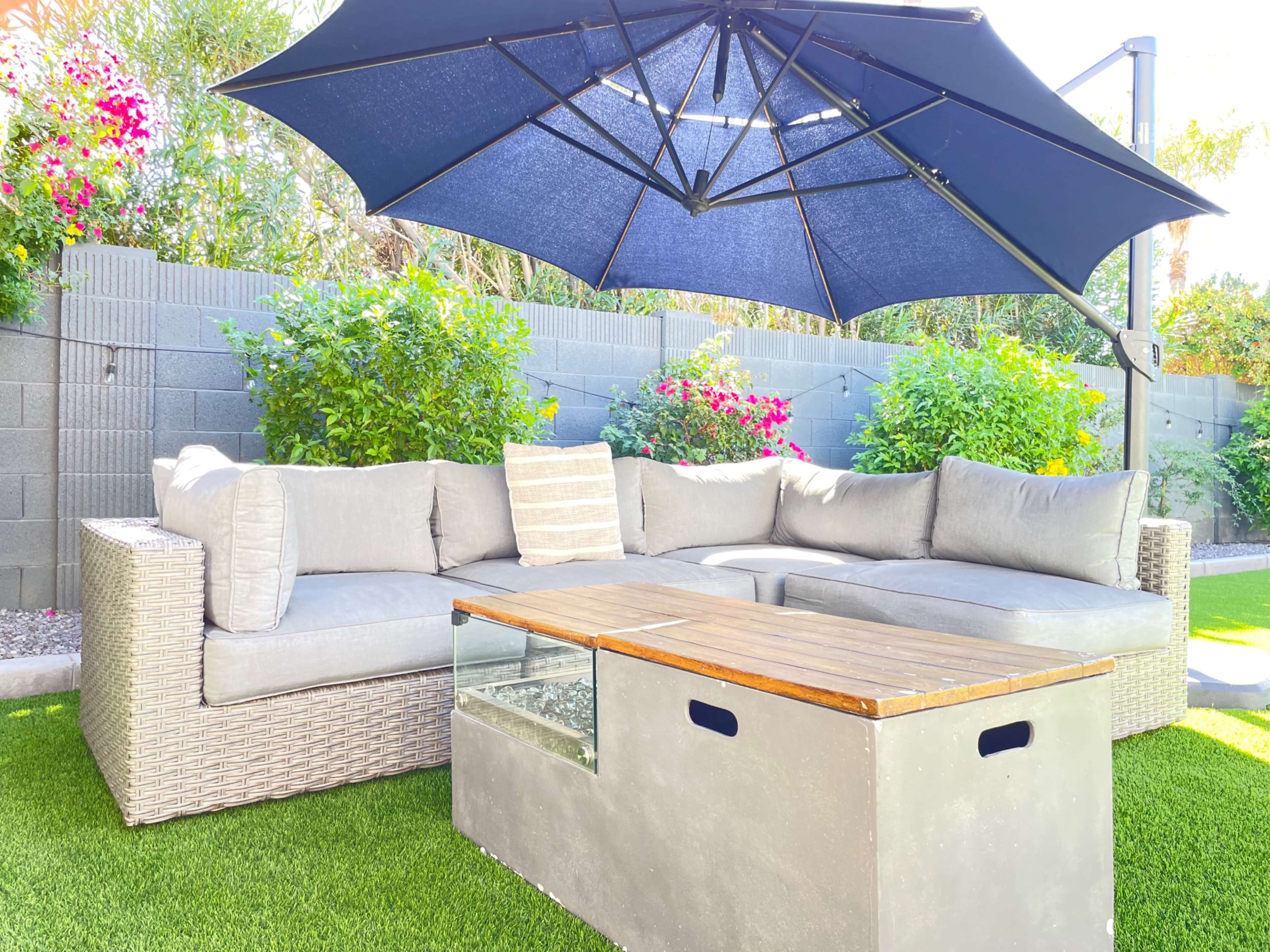 A sectional outdoor sofa with gray cushions is positioned under a large black umbrella in a garden surrounded by colorful flowers and green shrubs.