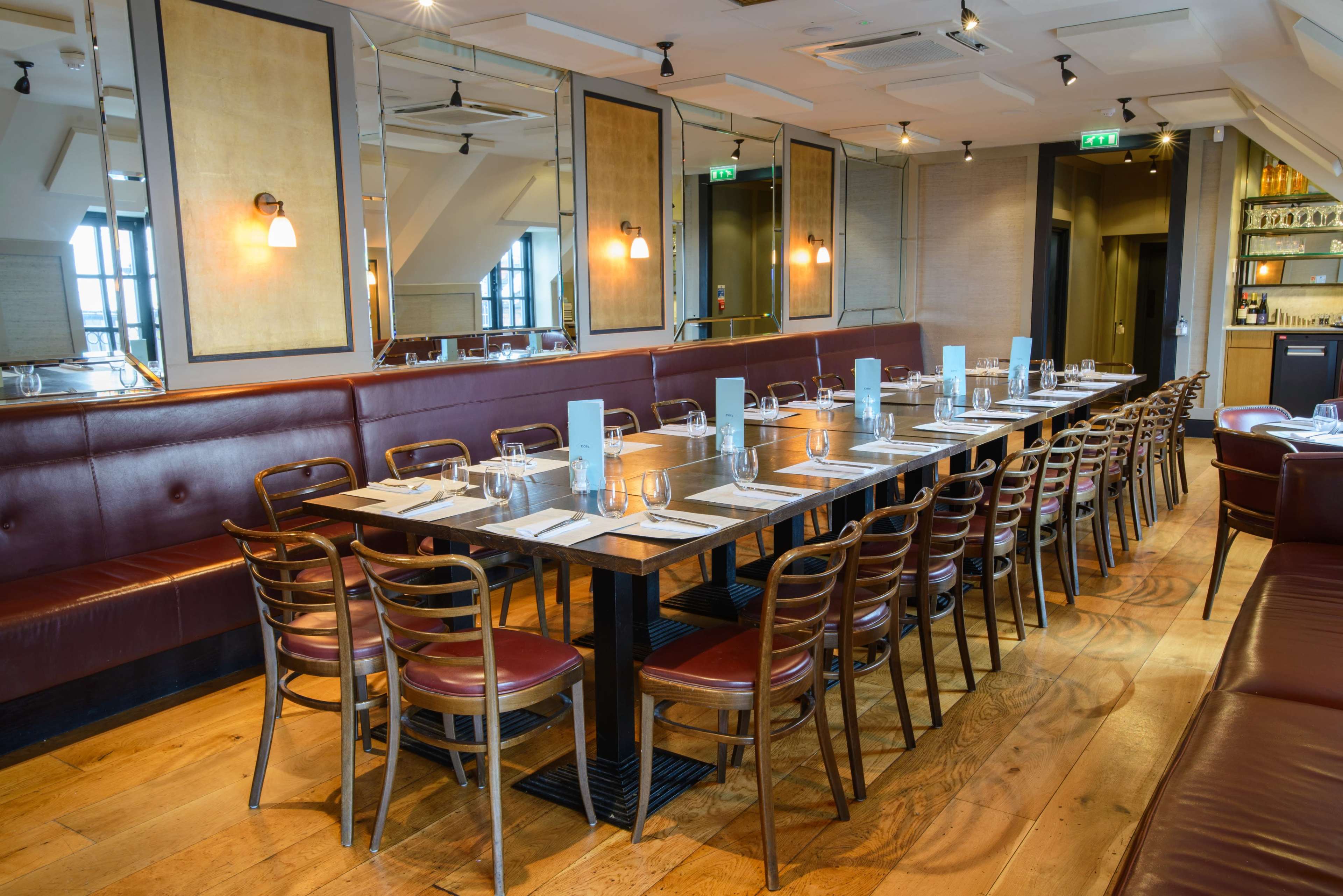A long wooden dining table is set with plates and glasses, surrounded by chairs in a modern restaurant with a mix of booths and wall-mounted lamps.