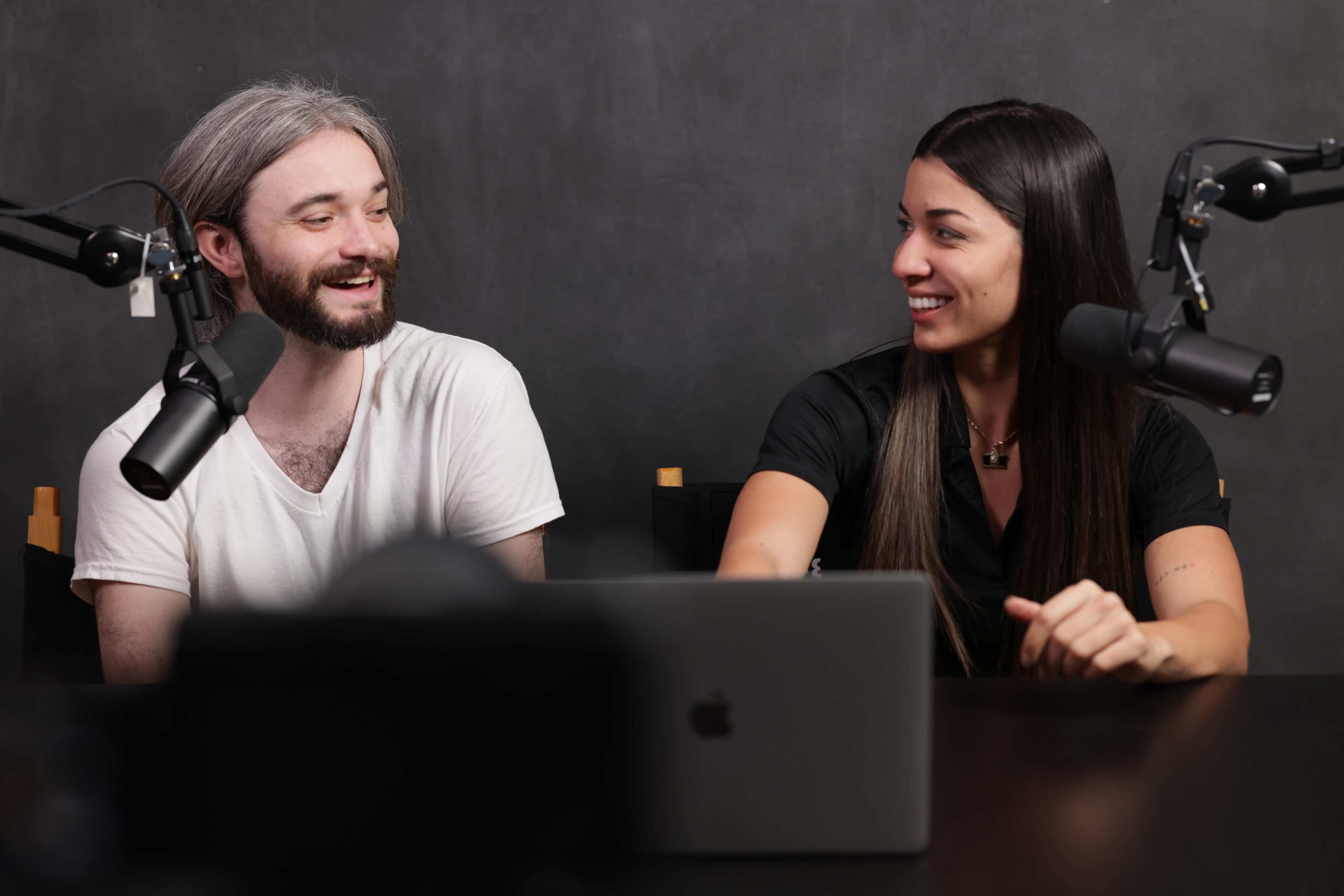 A man and a woman are smiling and sitting at a table with a laptop and microphones in a dark-colored studio setting.
