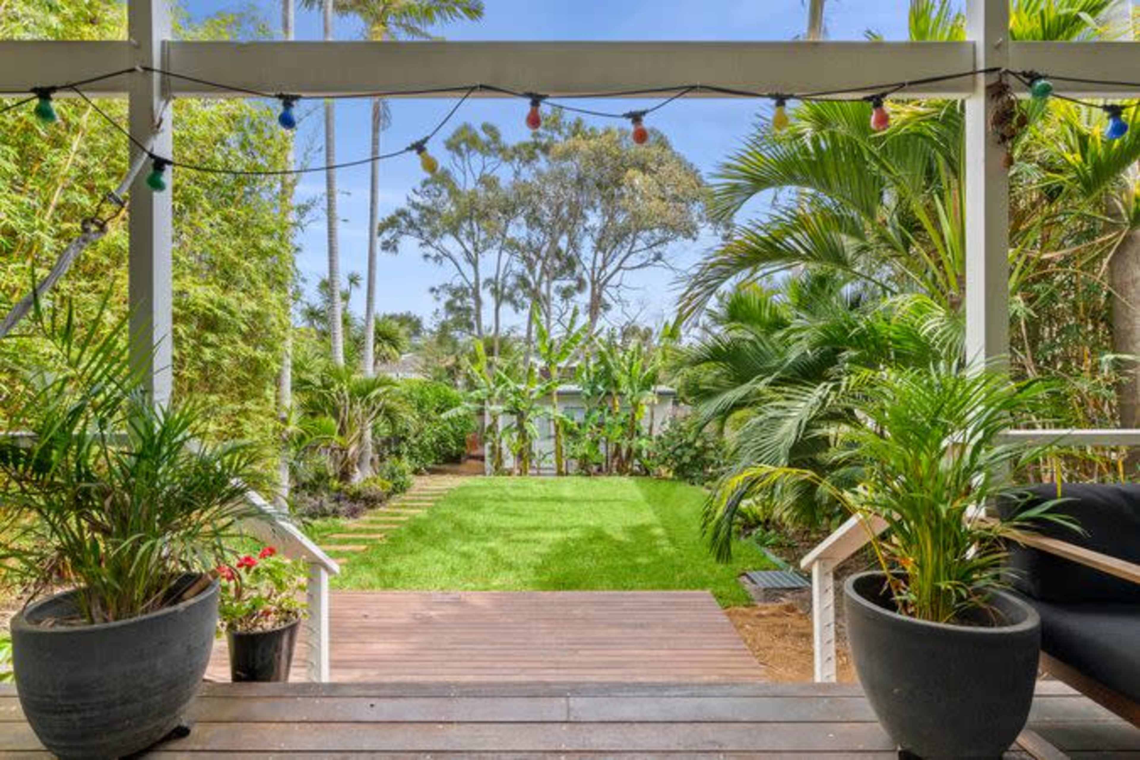 A wooden deck leads to a green lawn surrounded by lush plants and trees.