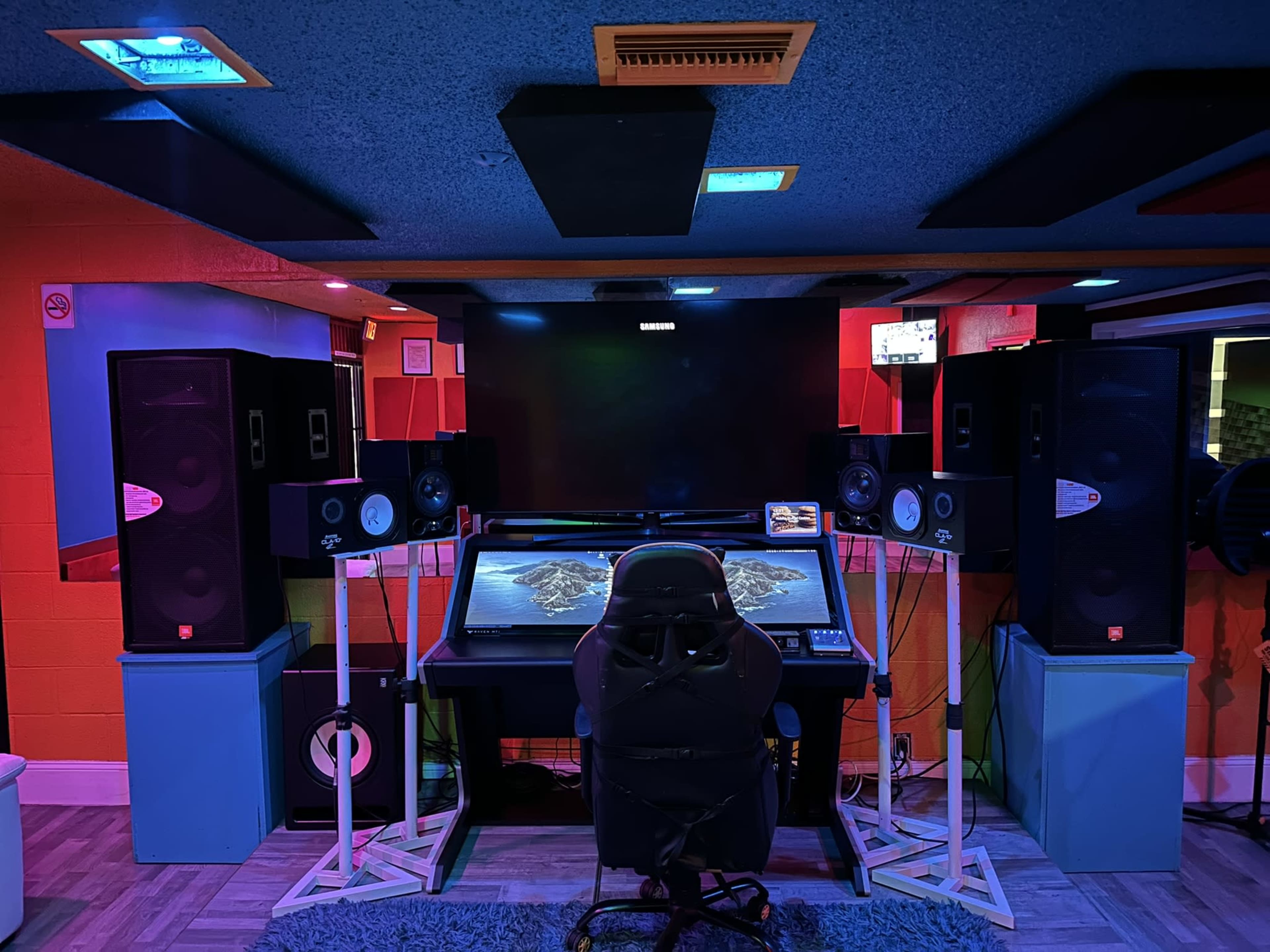A music studio with a central mixing desk, large speakers on either side, and a television mounted above.