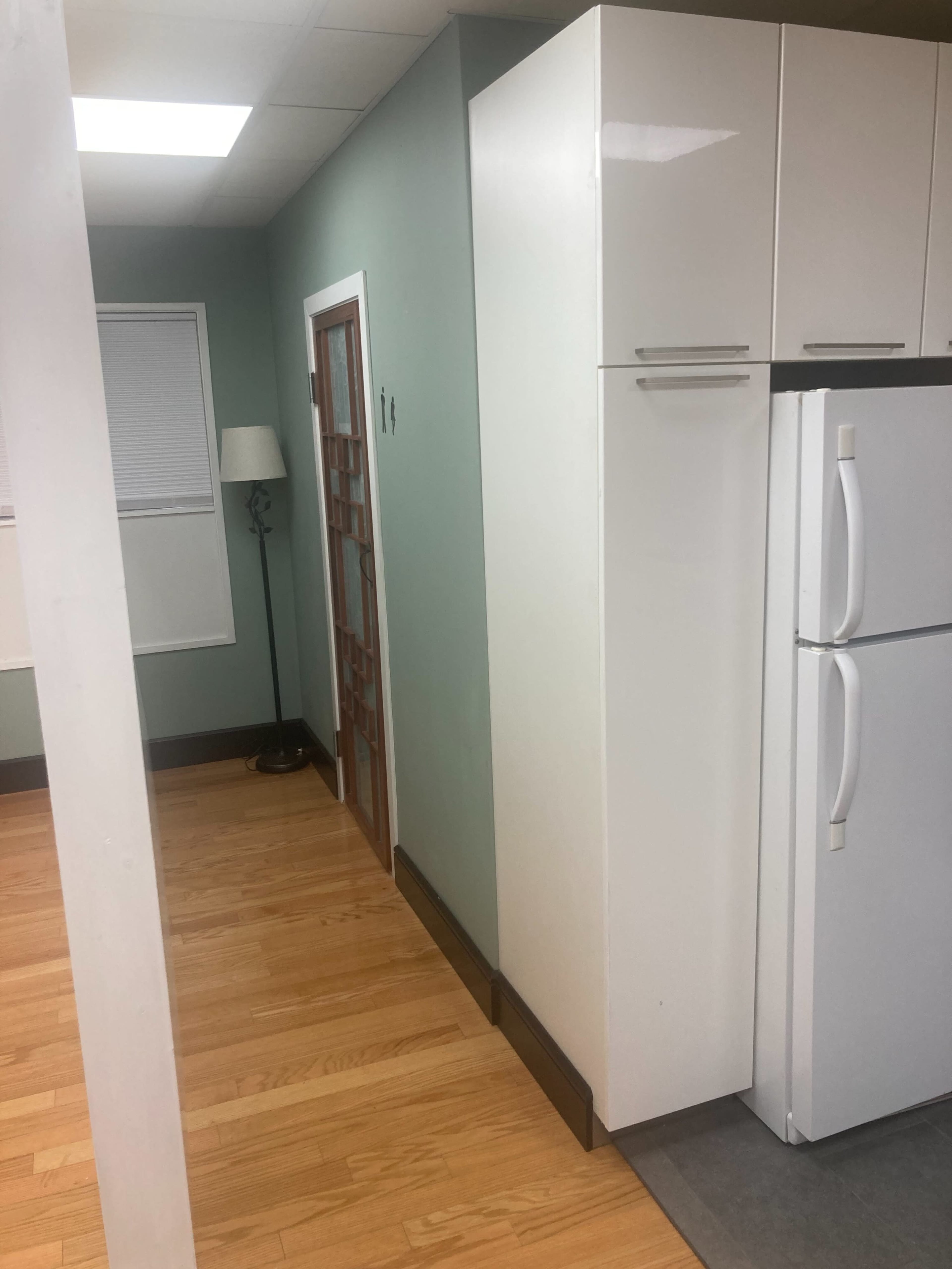 A kitchen area with a white refrigerator, a tall white cabinet, and a light green wall beside a door leading to another room.