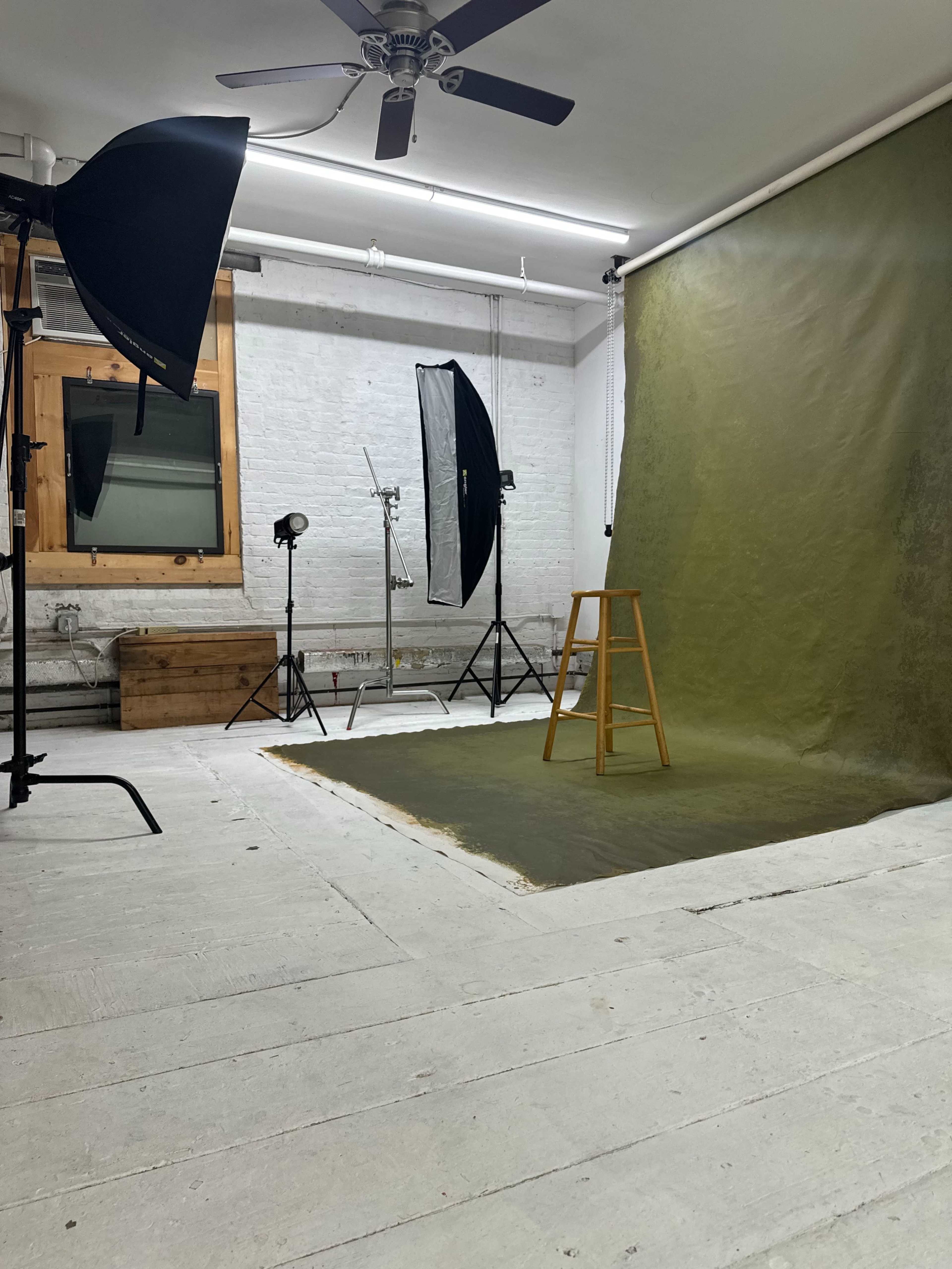 The image shows a photography studio featuring a green backdrop, a wooden stool, and various lighting equipment.