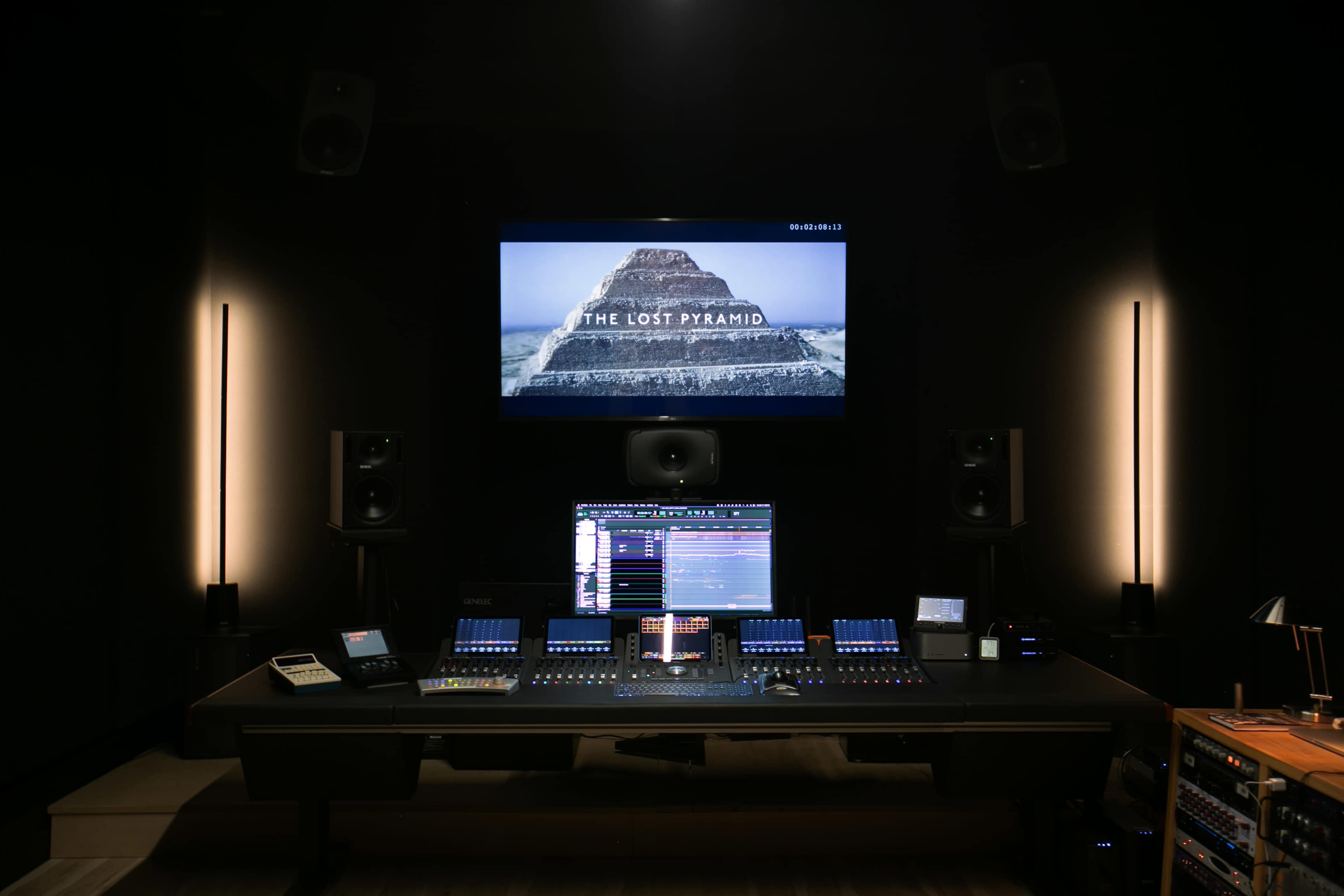 A modern audio mixing studio with a large screen displaying "THE LOST PYRAMID" and an array of digital mixing equipment on the central desk.