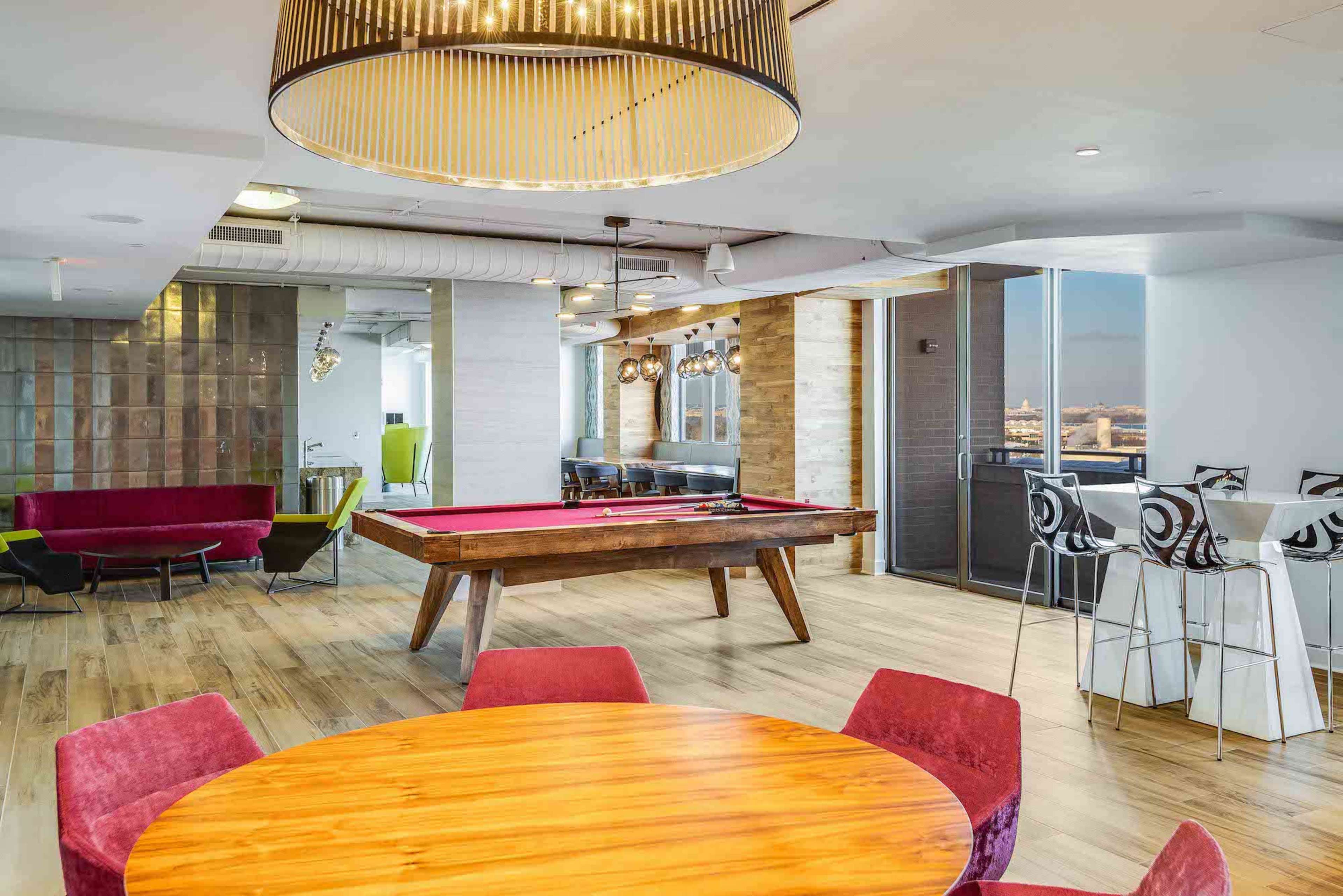 A spacious lounge area with a wooden pool table, a round dining table surrounded by pink chairs, and a bar counter with high stools, all set in a modern office environment.