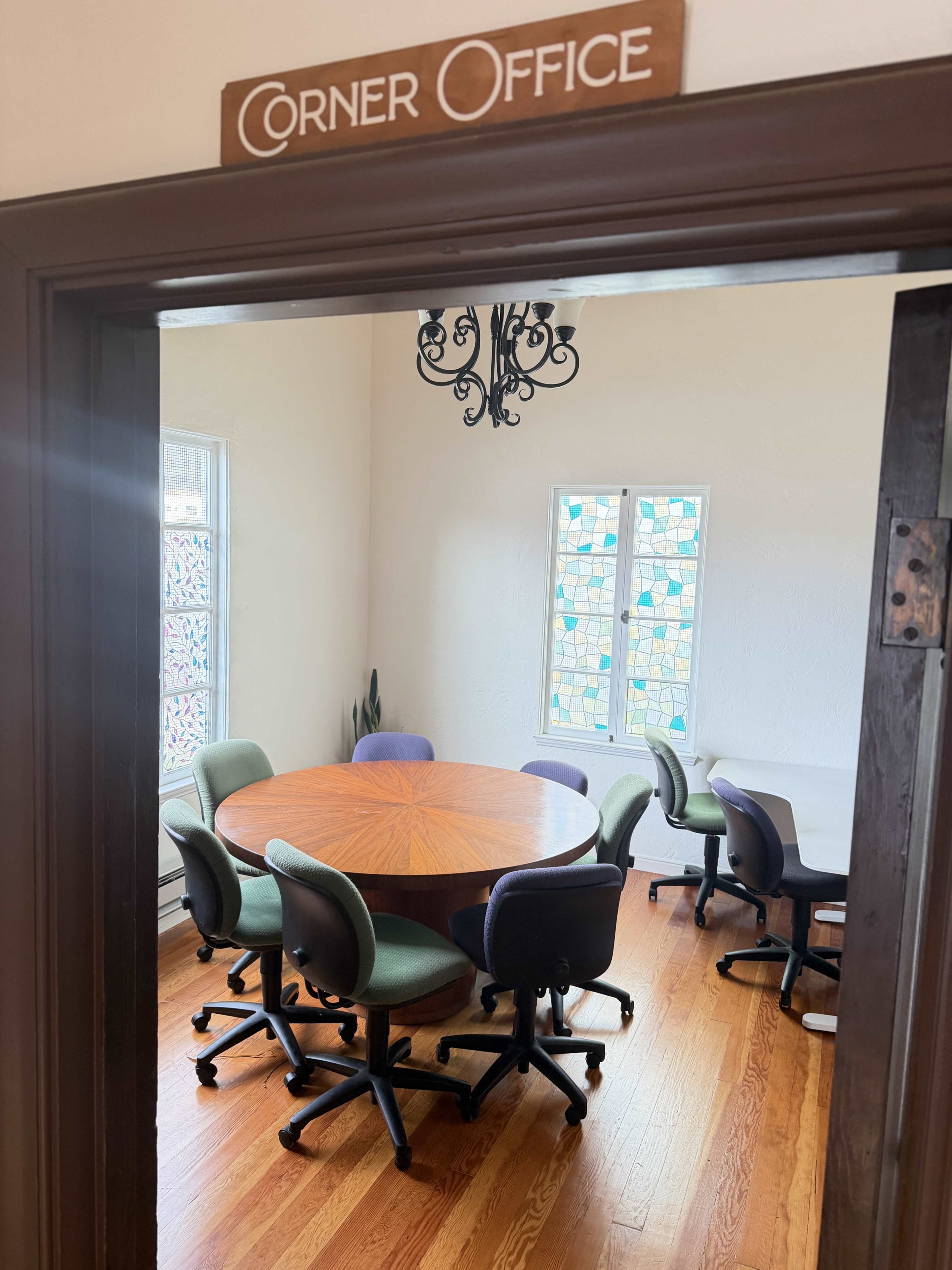 The image shows a corner office featuring a round wooden table surrounded by several office chairs and two stained glass windows.