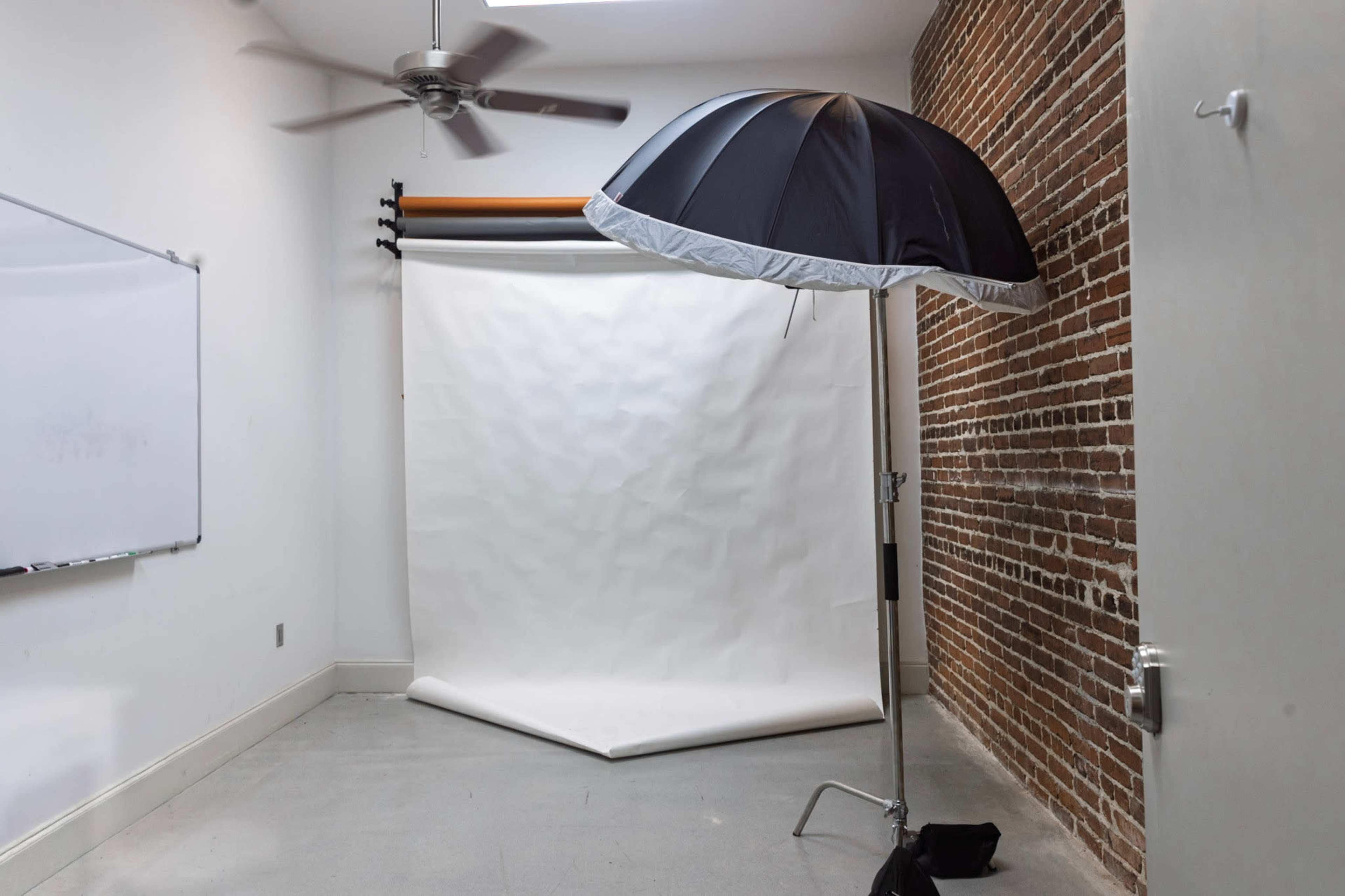 The image shows a photography studio with a white backdrop, a large black umbrella reflector, and a brick wall.
