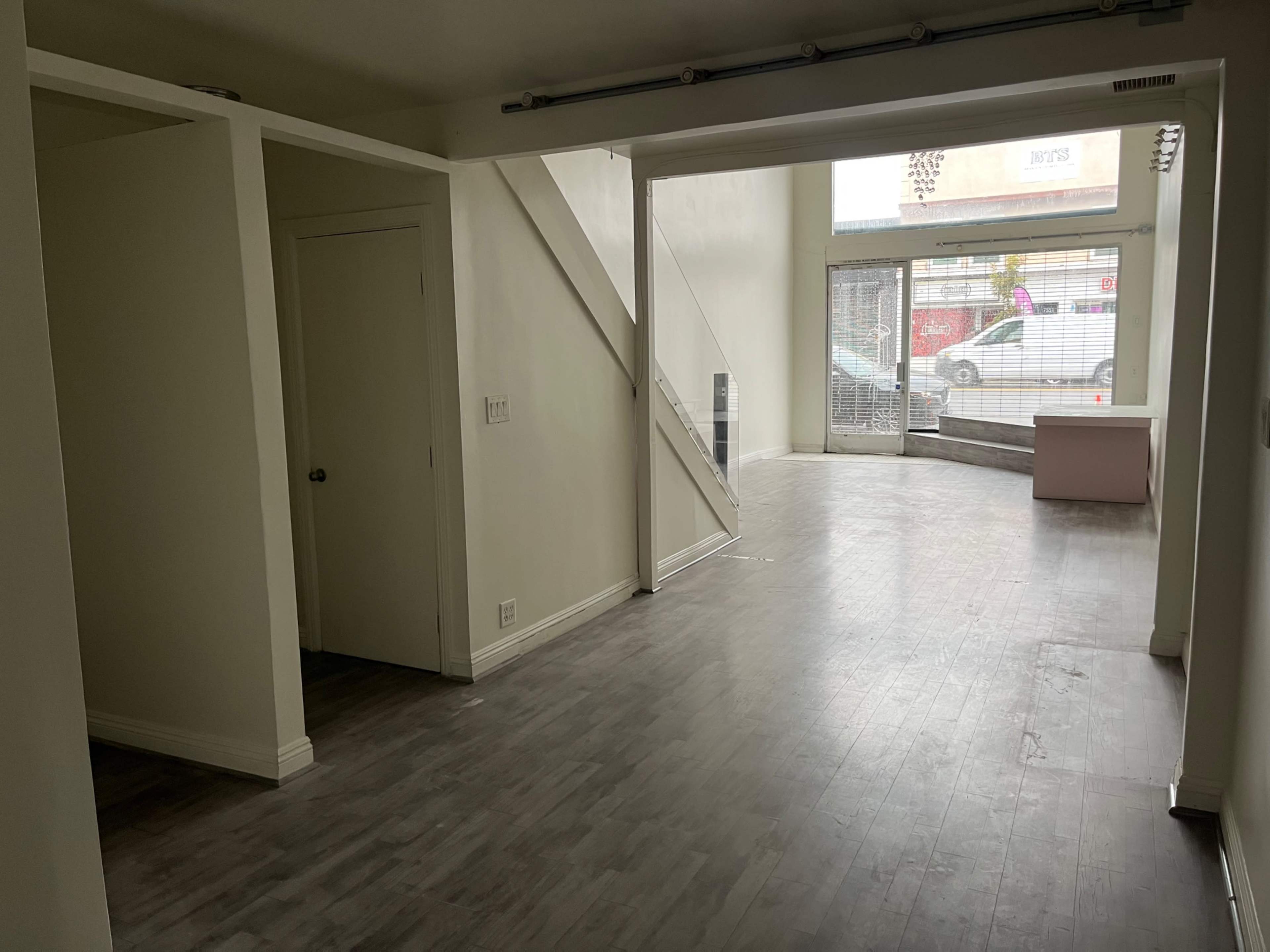 The image shows a spacious, empty room featuring gray laminate flooring, a staircase leading to an upper level, and large windows at the far end that provide natural light.