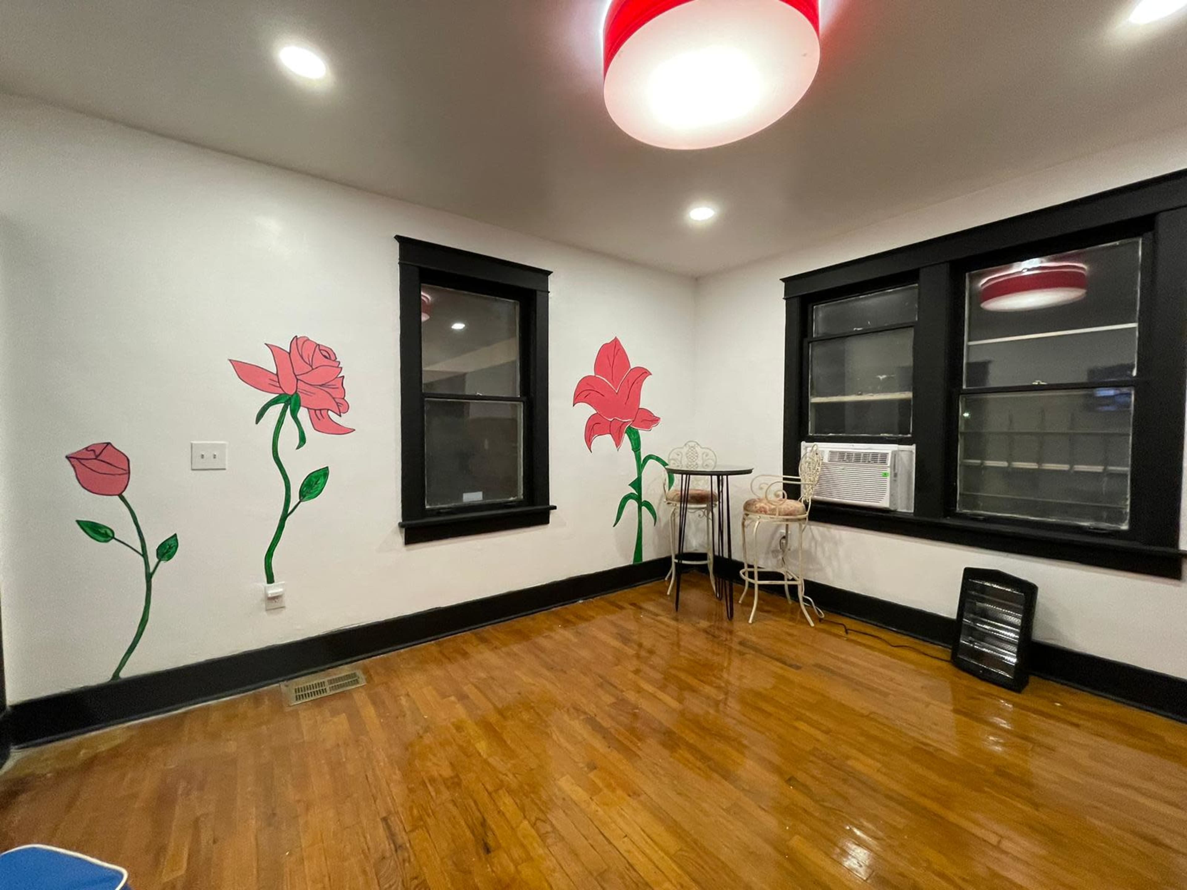 The room features hardwood floors, black window frames, and painted flower murals on the white walls, along with a small table and an air conditioning unit.