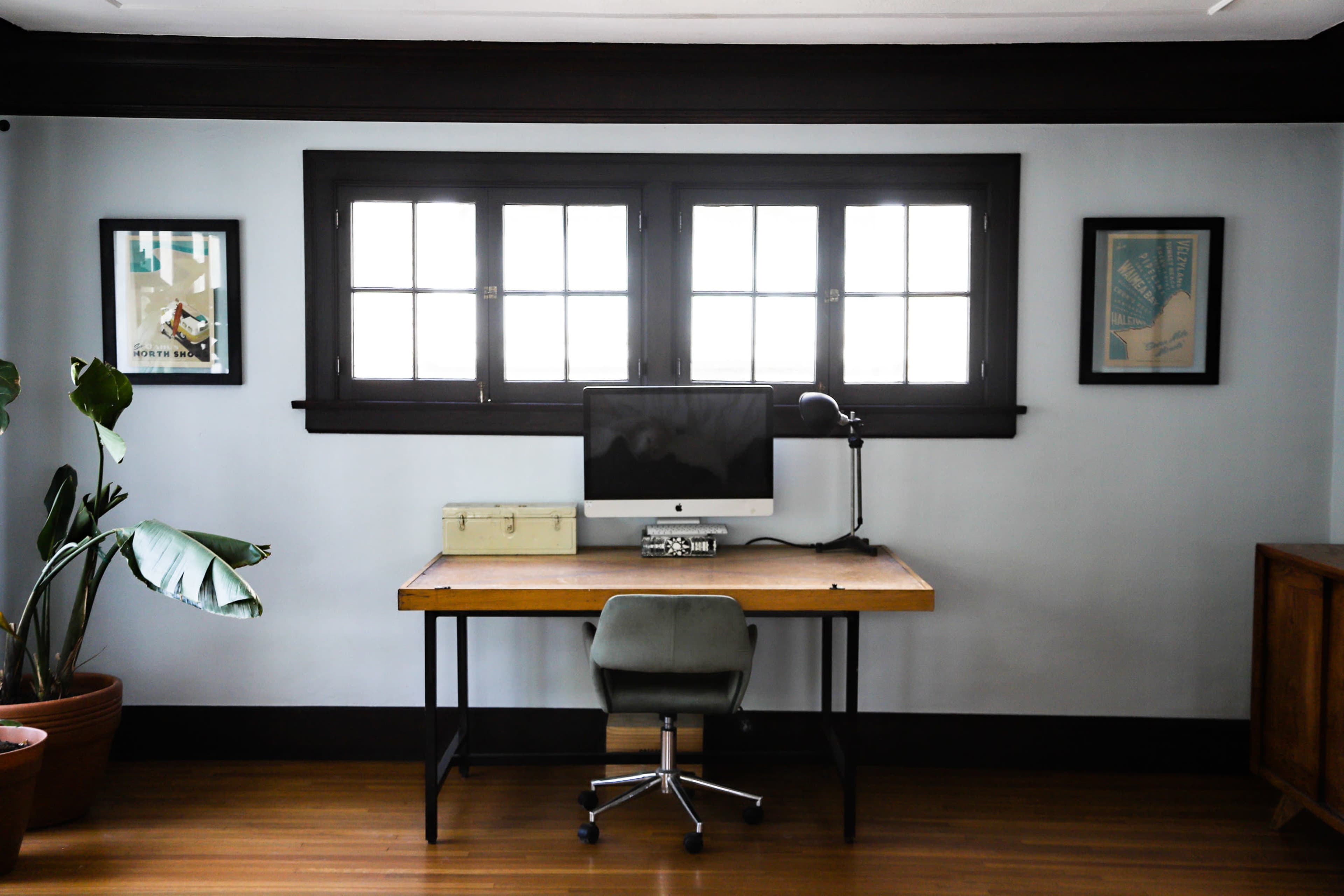 A desk with an iMac computer sits in front of a window flanked by framed artwork, with a potted plant in the corner of the room.