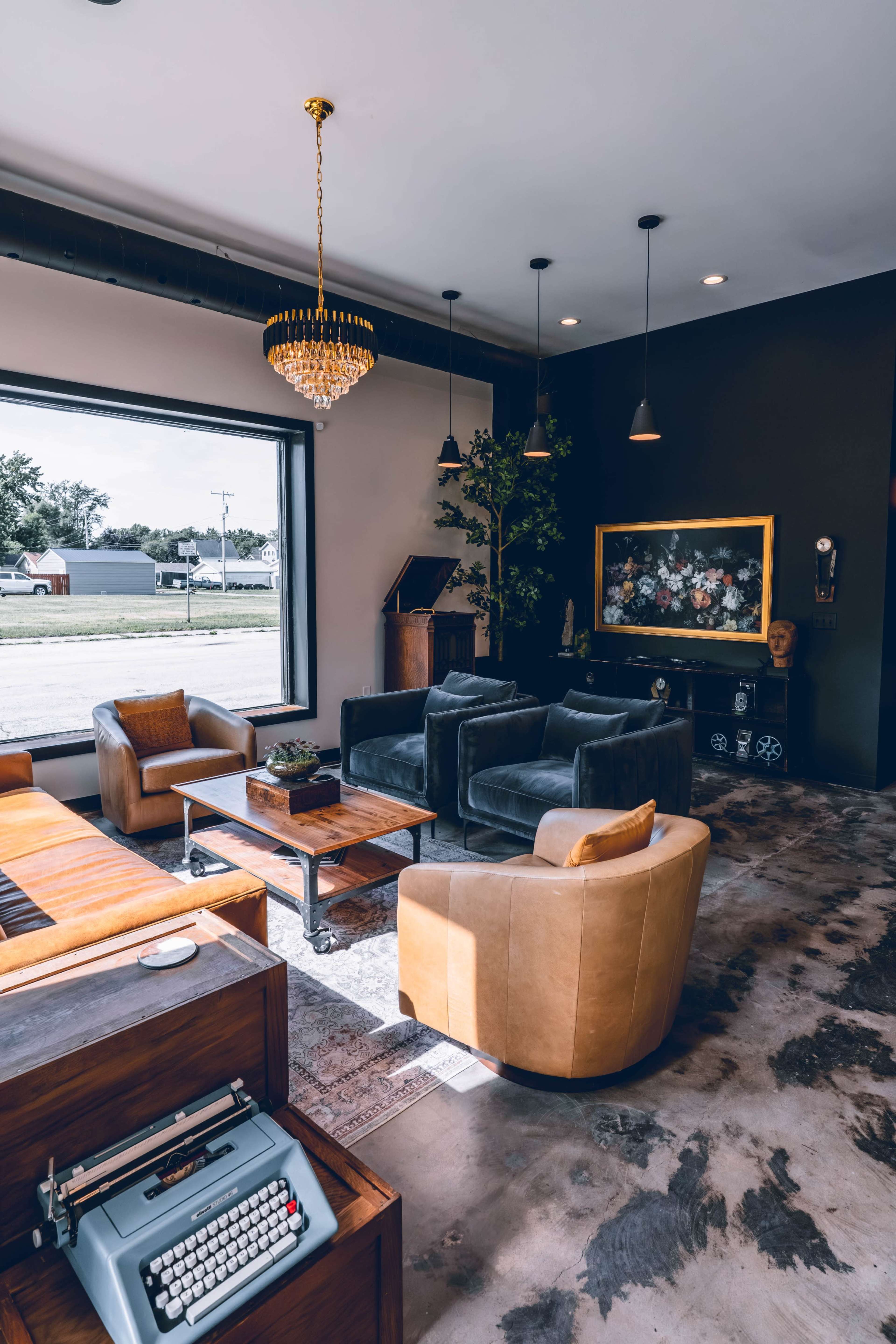 The image shows a modern lounge area with comfortable seating, a coffee table, a typewriter, and decorative plants, all set against large windows allowing natural light to enter.