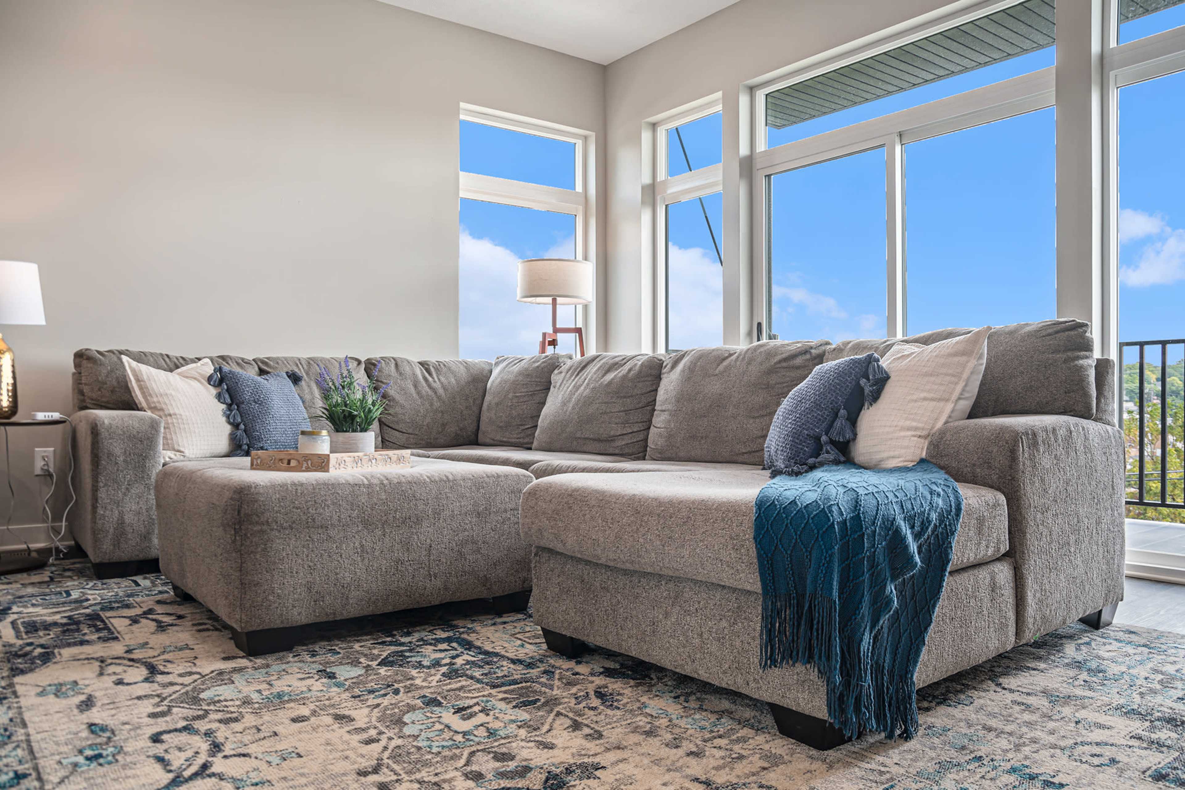 A large gray sectional sofa with decorative pillows sits on a patterned rug in a bright room with large windows overlooking a blue sky.