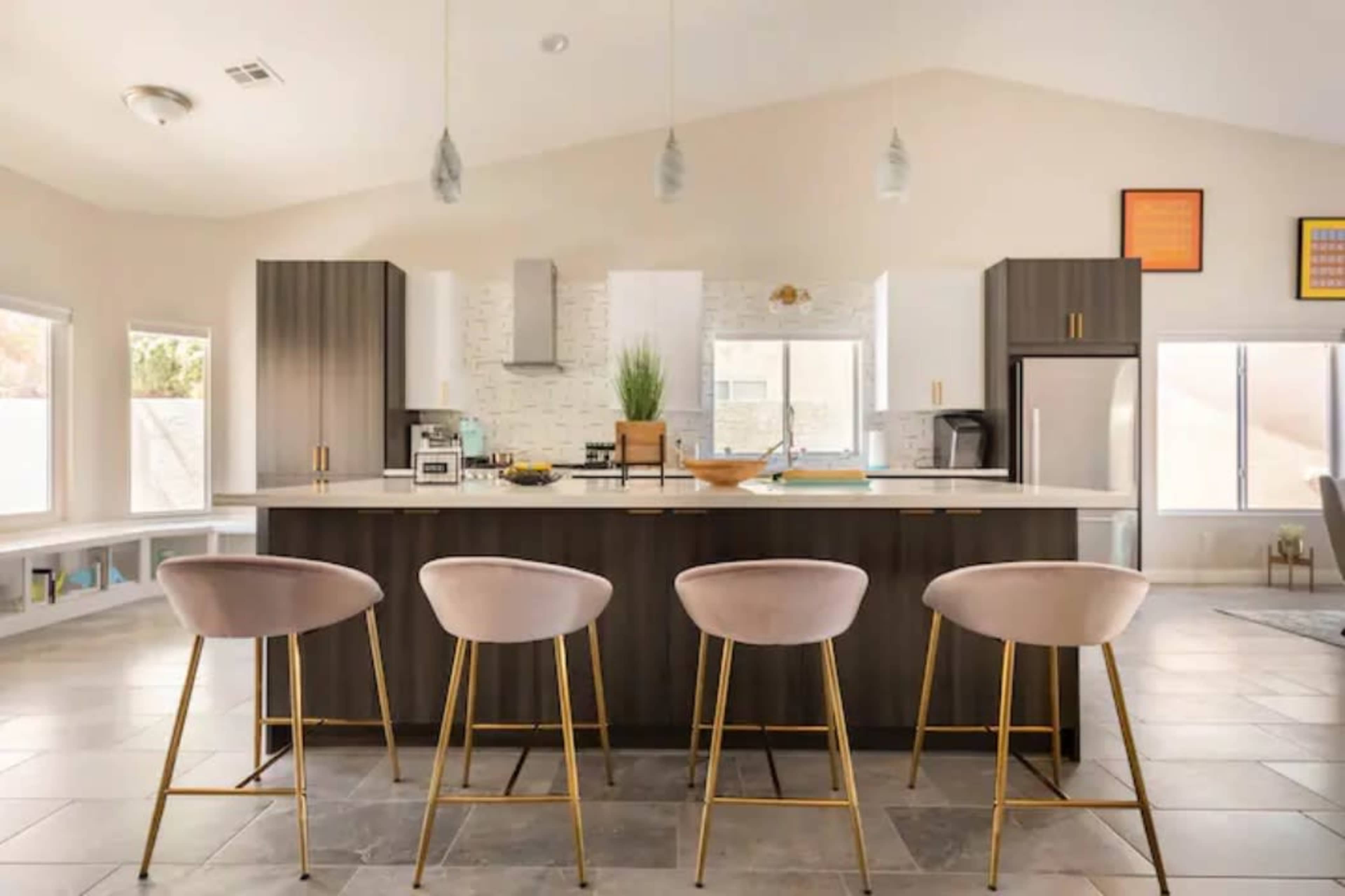 A modern kitchen features a central island with four upholstered bar stools, surrounded by sleek cabinetry and large windows.