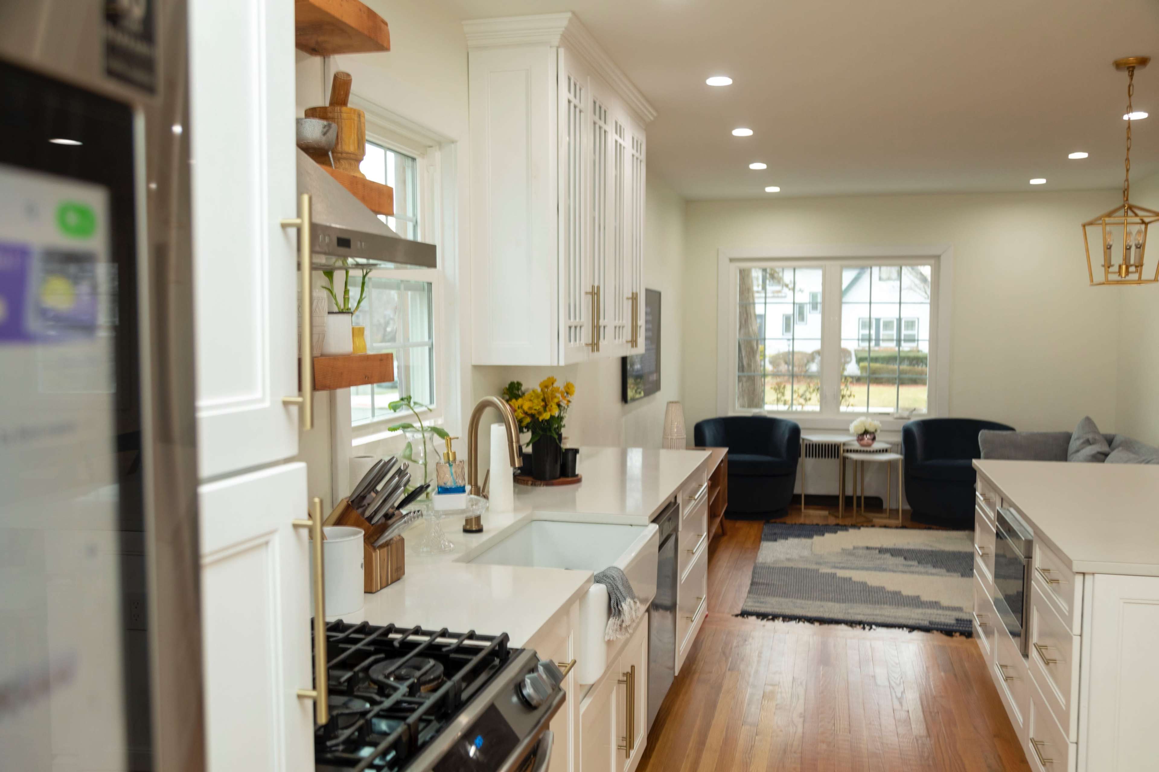 The image depicts a modern kitchen with white cabinetry, a central island, and a view of a connected living area featuring two armchairs and a large window.