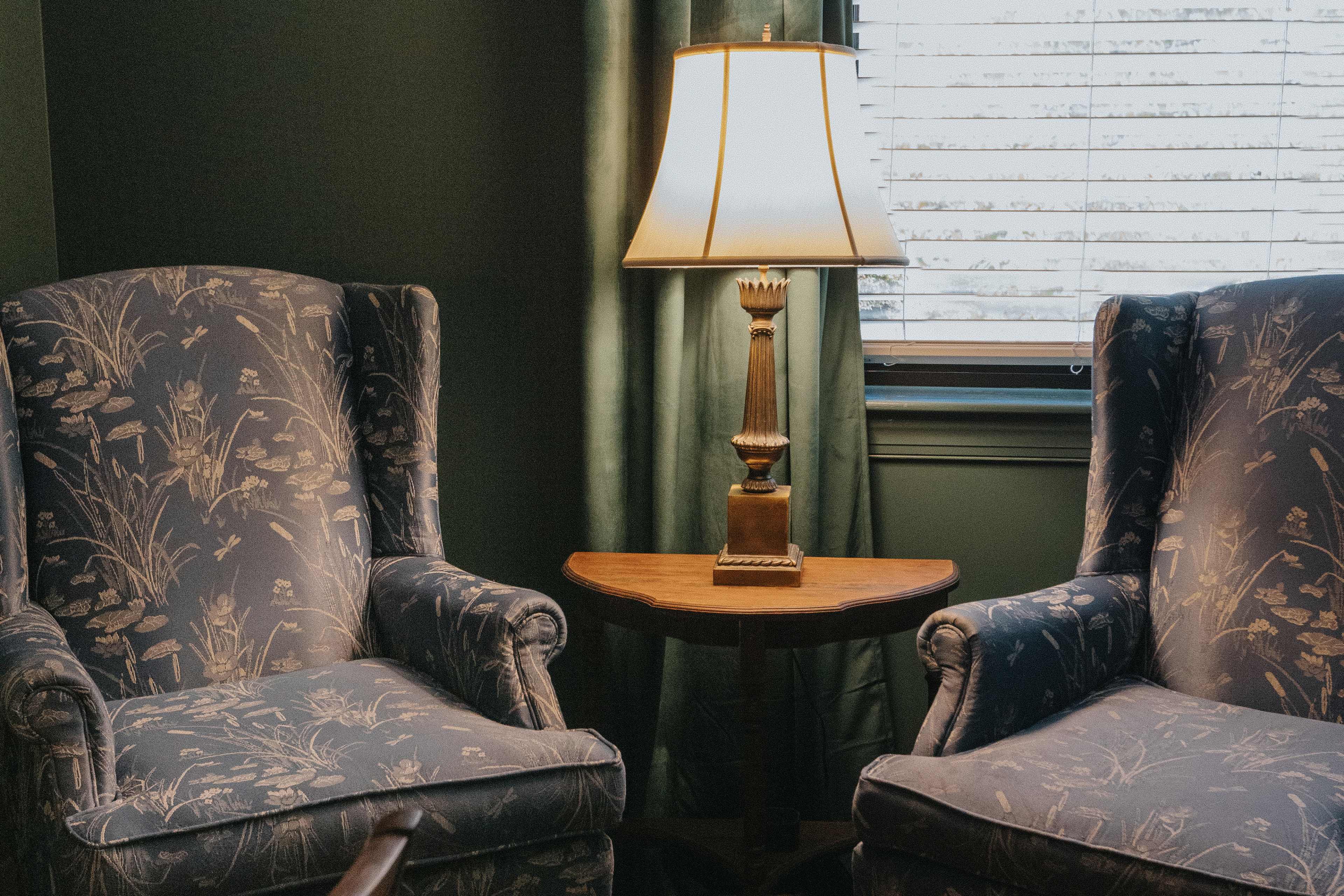 Two upholstered armchairs facing a small wooden table beside a lamp in a room with green walls and a window with blinds.