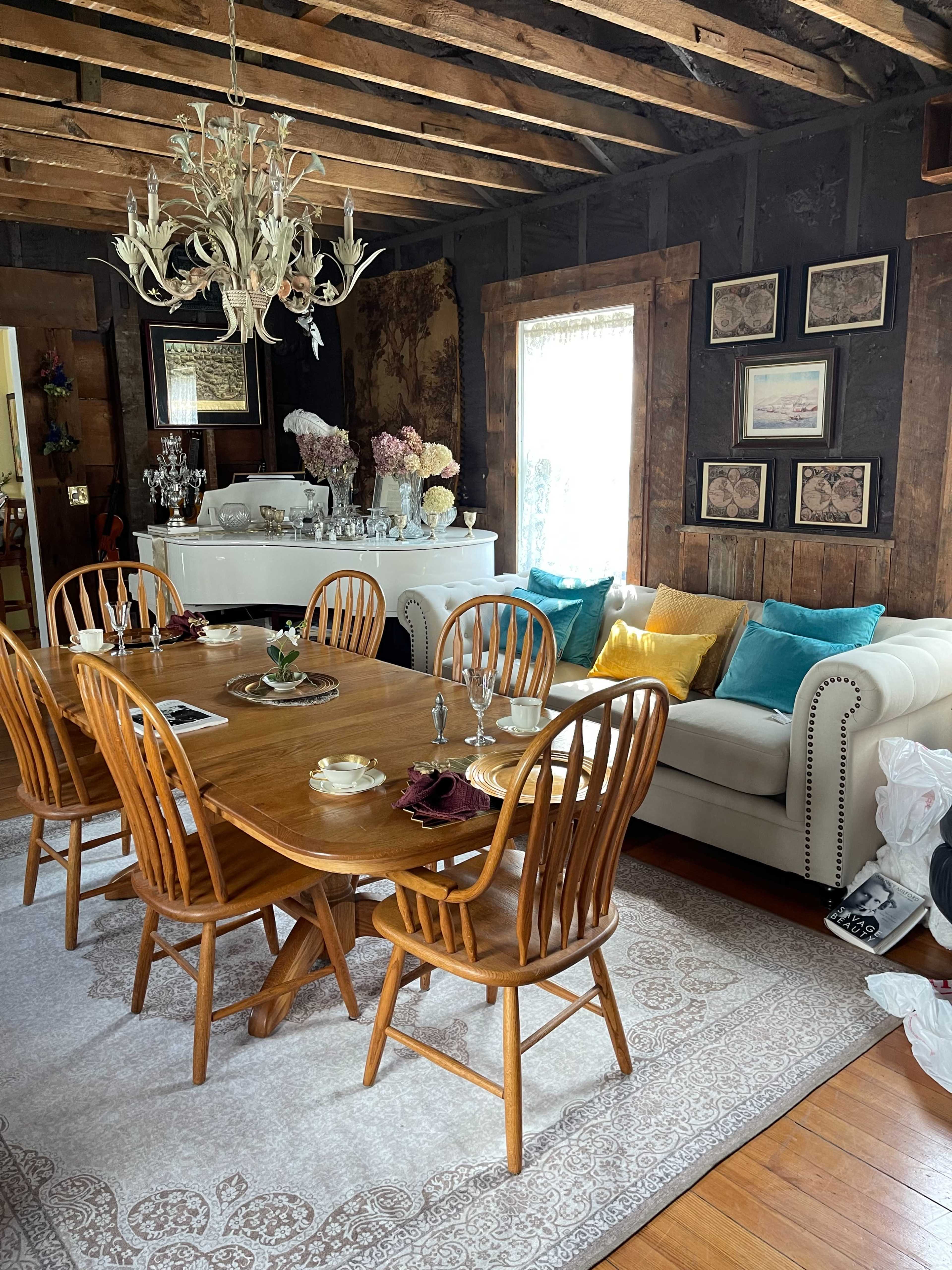 A wooden dining table surrounded by chairs is set in a rustic room with a white couch, decorative pillows, and a chandelier overhead.