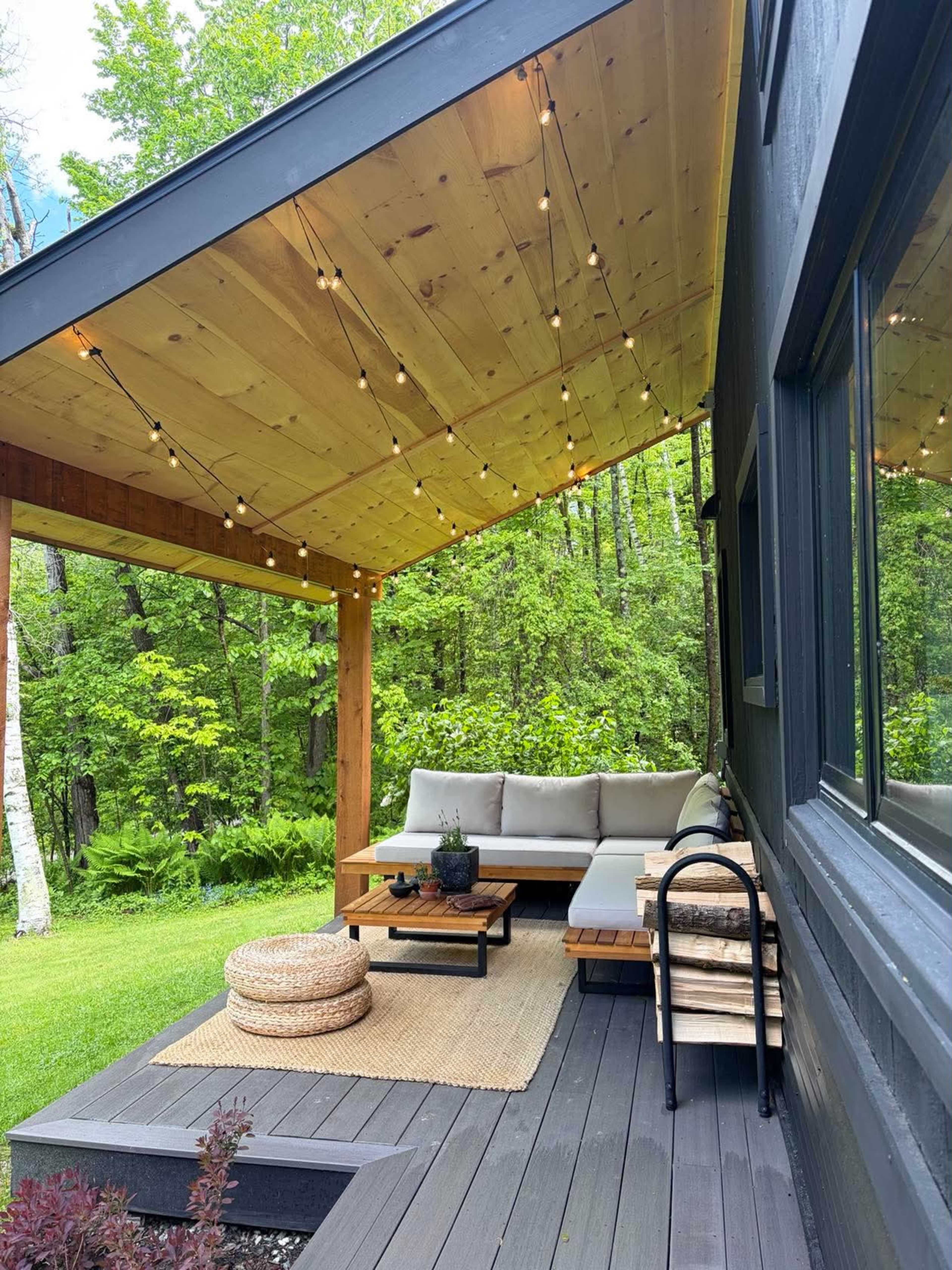 The image features a cozy outdoor seating area on a wooden deck covered by a sloped roof, adorned with string lights and surrounded by lush greenery.