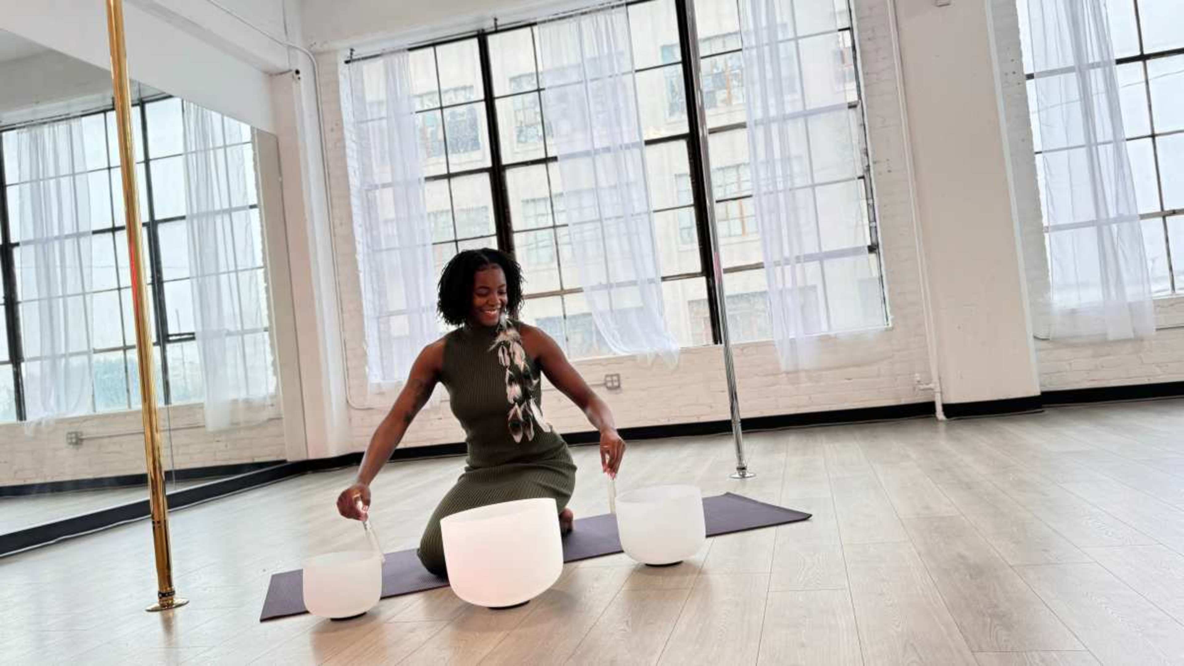 A person sits on a yoga mat in a bright studio, arranging three crystal sound bowls in front of them.