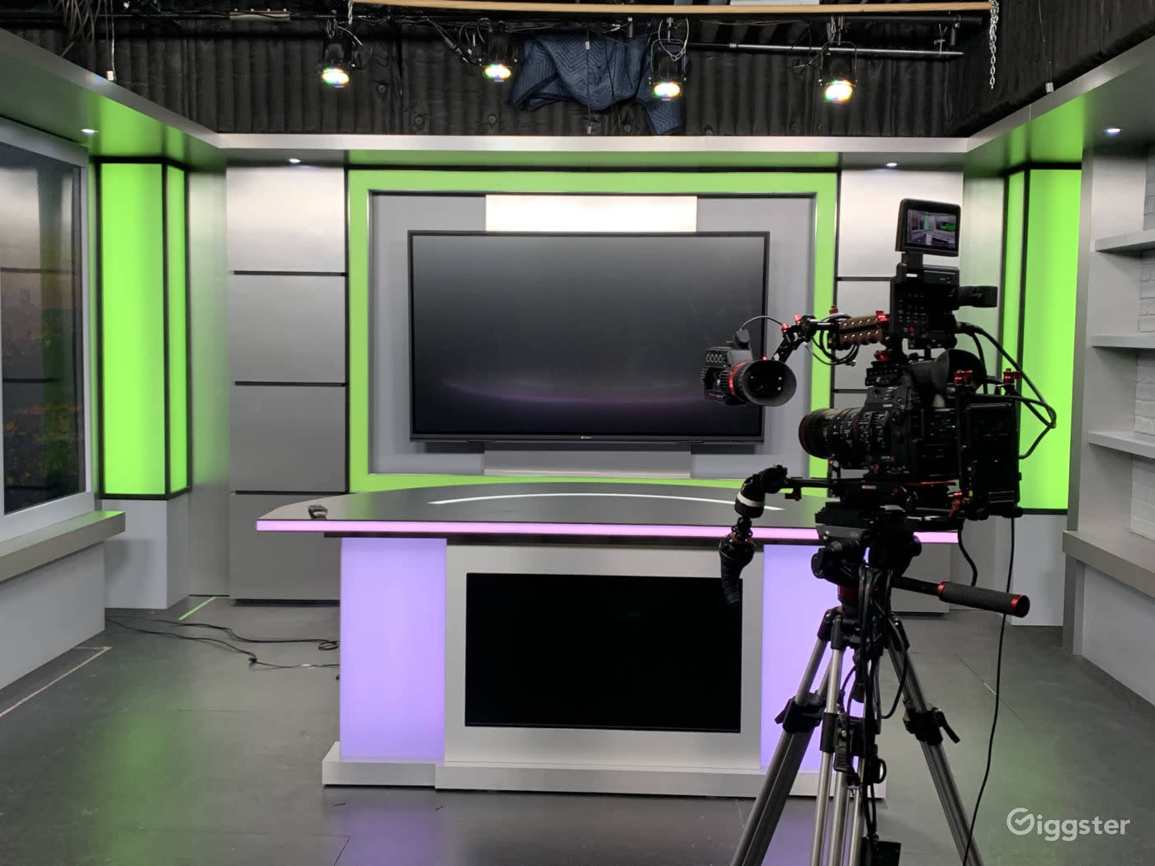 A modern television studio setup with a large screen and a camera positioned on a tripod, illuminated by green and purple lighting.