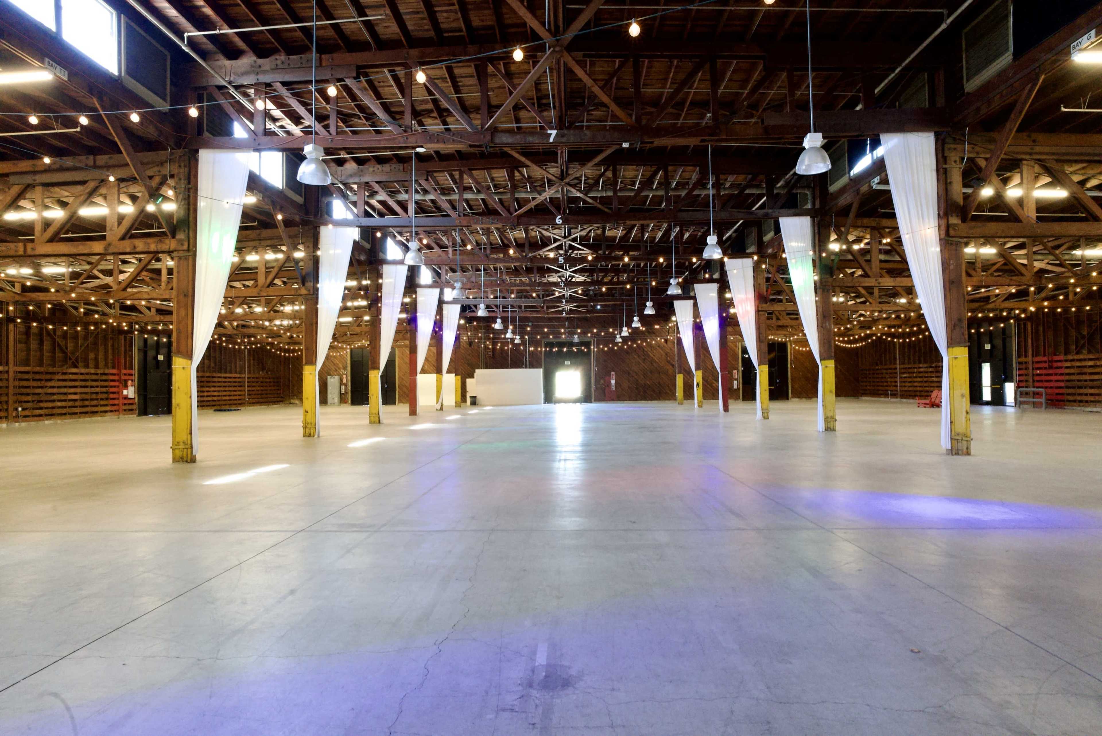 The image shows a spacious, well-lit warehouse interior with exposed wooden beams, hanging lights, and colorful lighting on the concrete floor.
