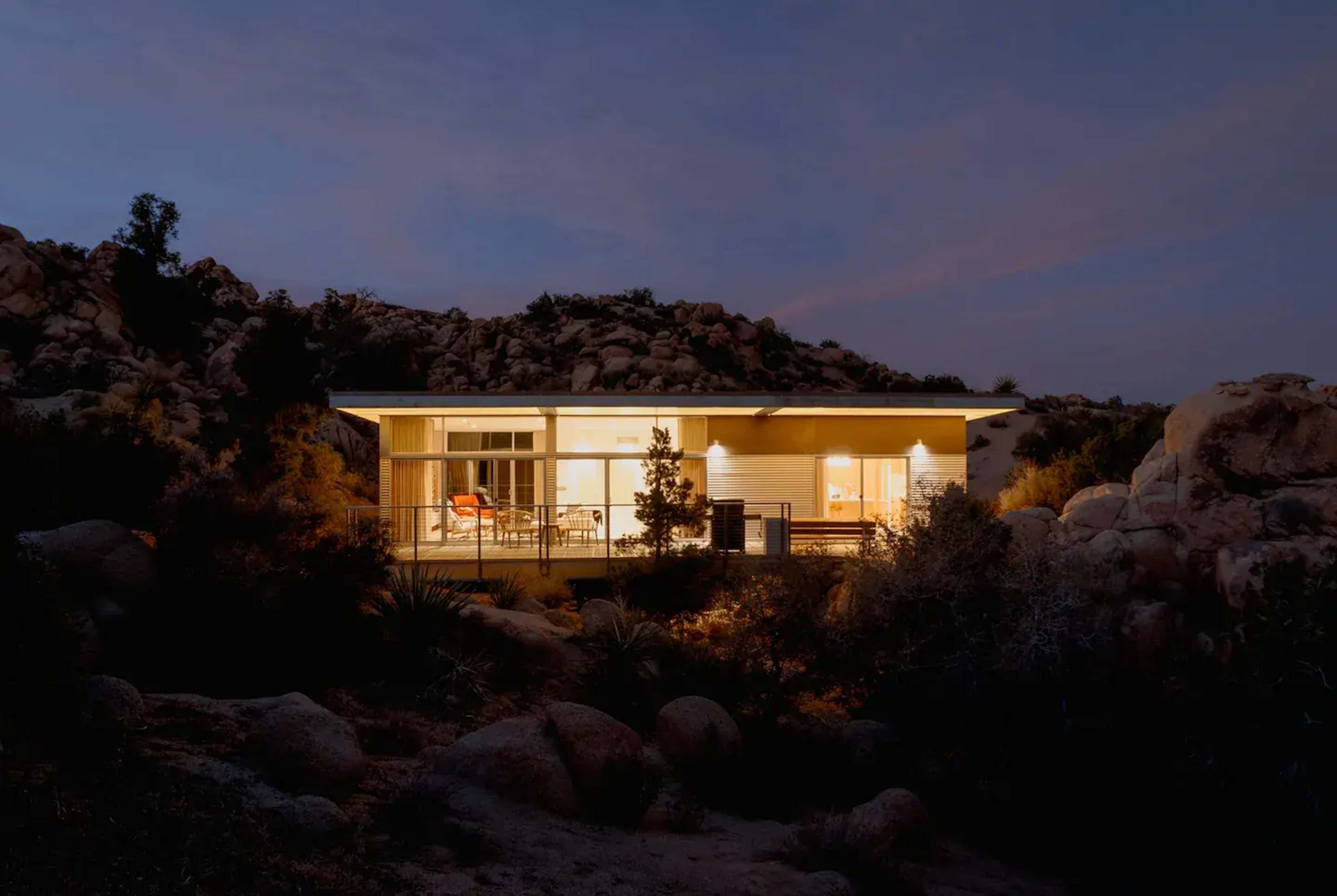 A modern house with large windows is illuminated at dusk, situated among rocky terrain and desert vegetation.