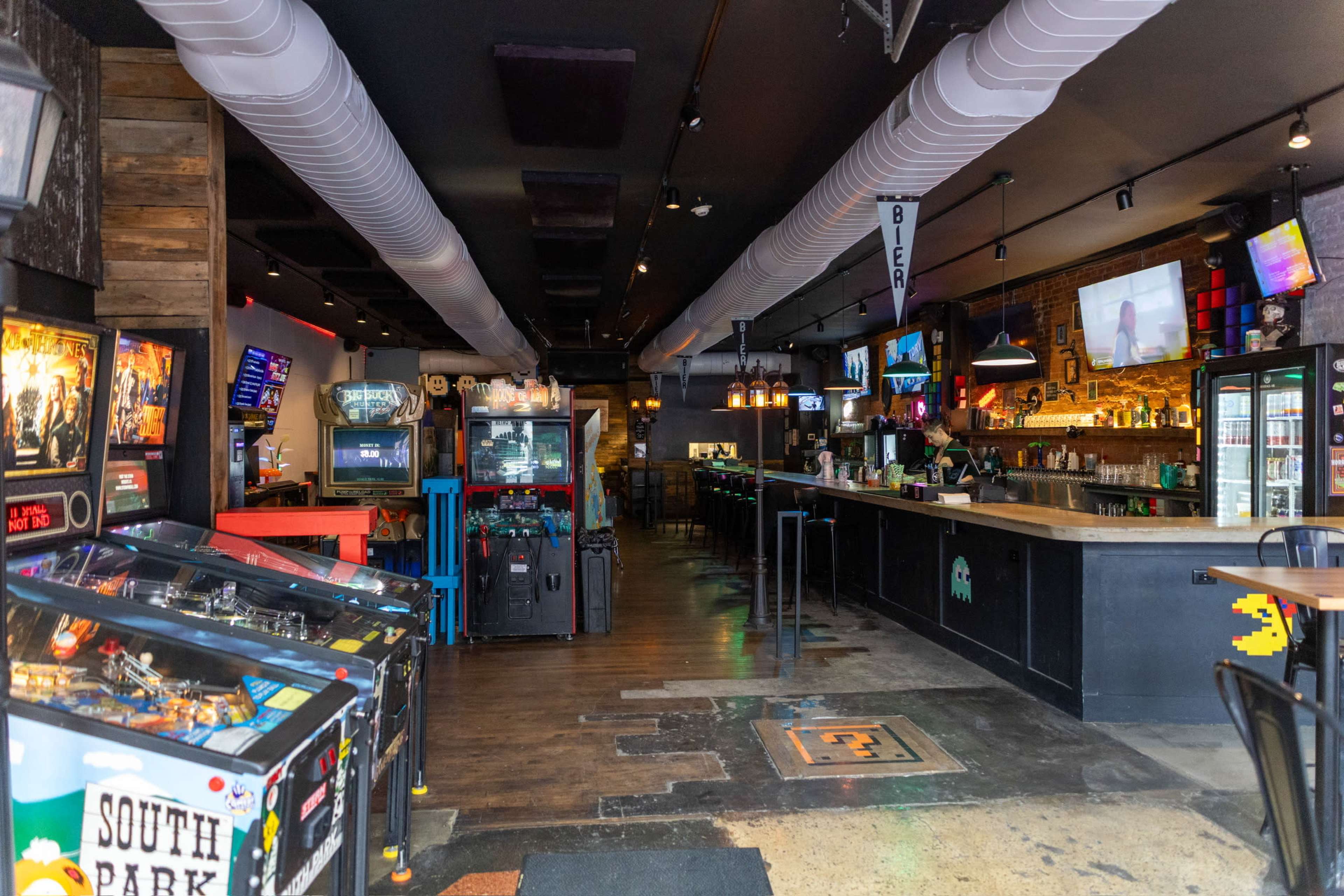 The image shows a dimly lit bar and arcade space featuring several arcade machines, a long wooden bar, and various gaming memorabilia on the walls.