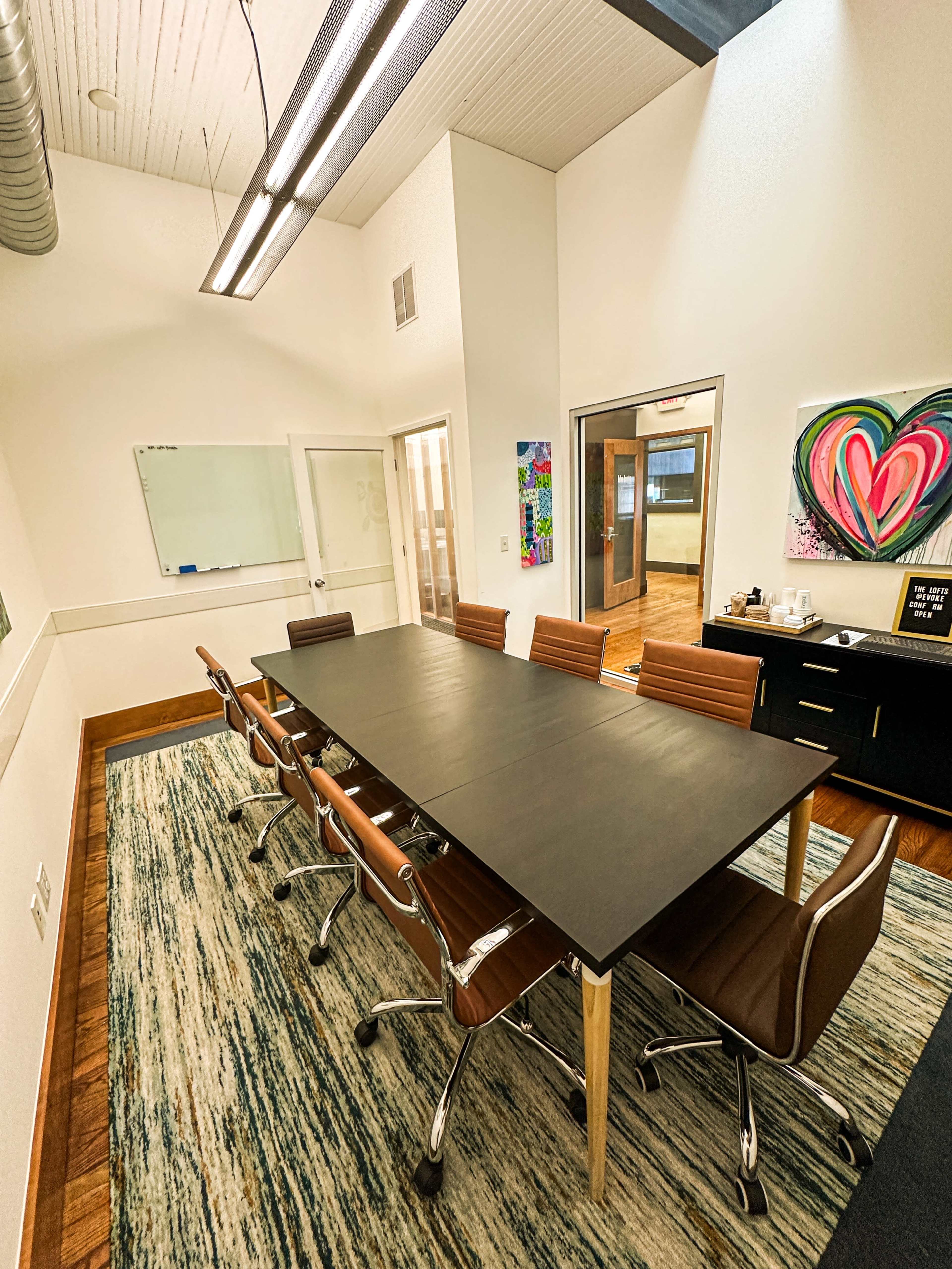 A modern conference room features a long black table surrounded by brown leather chairs on a patterned rug, with a whiteboard and colorful artwork on the walls.