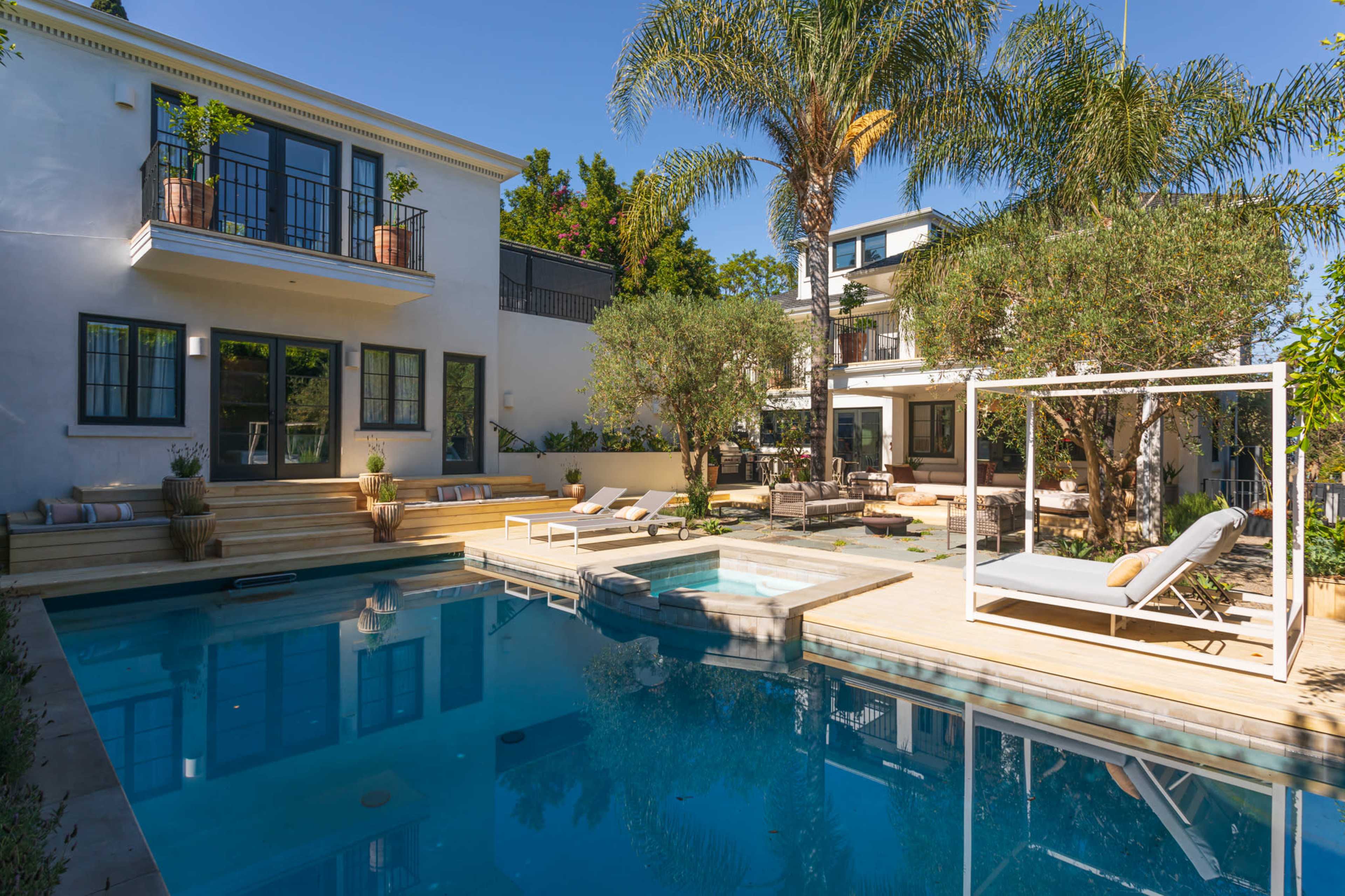 A modern backyard features a swimming pool, spa, and lounge area surrounded by palm trees and landscaped gardens.