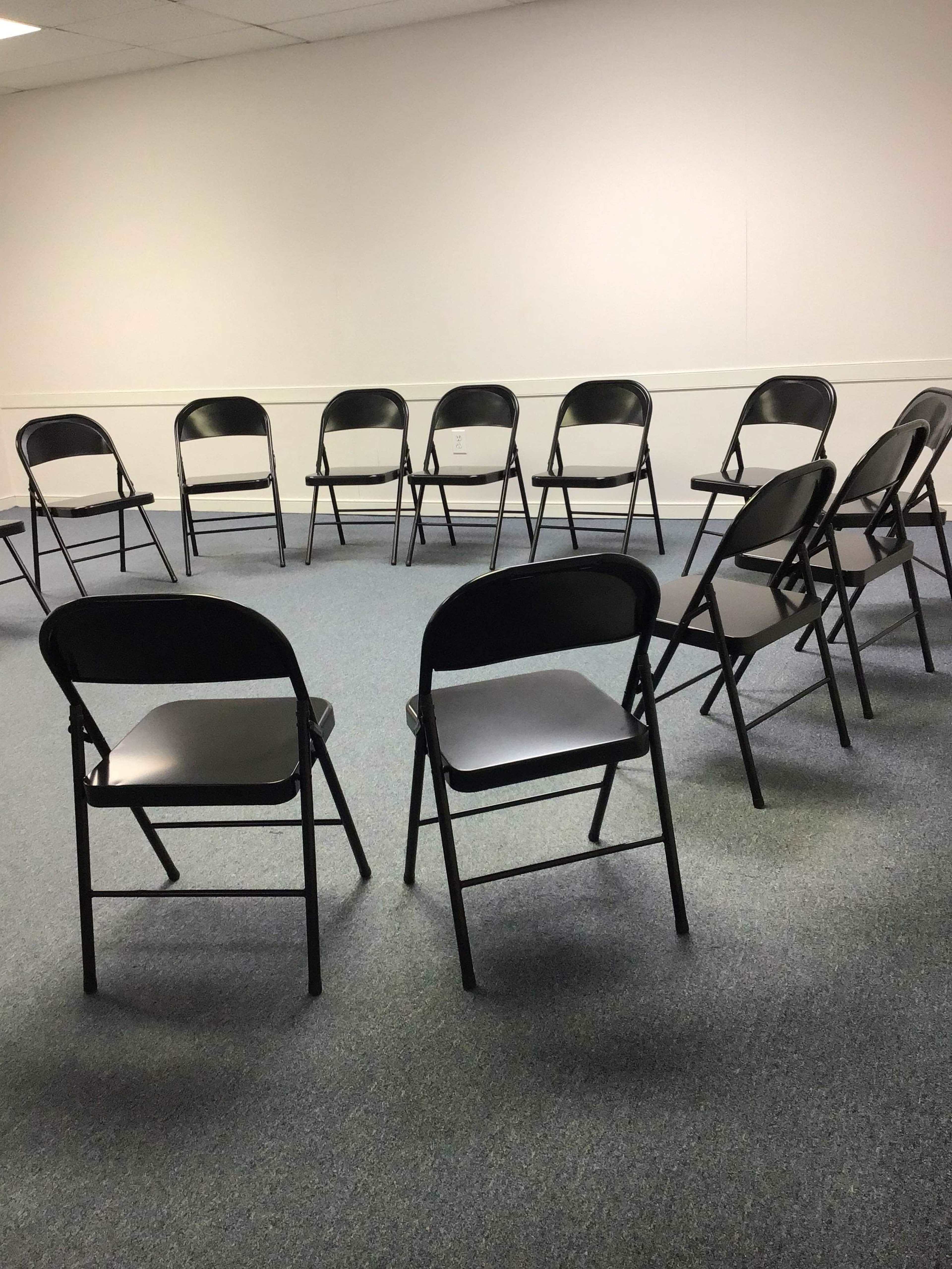 A circle of folding chairs is arranged on a carpeted floor in a plain room.