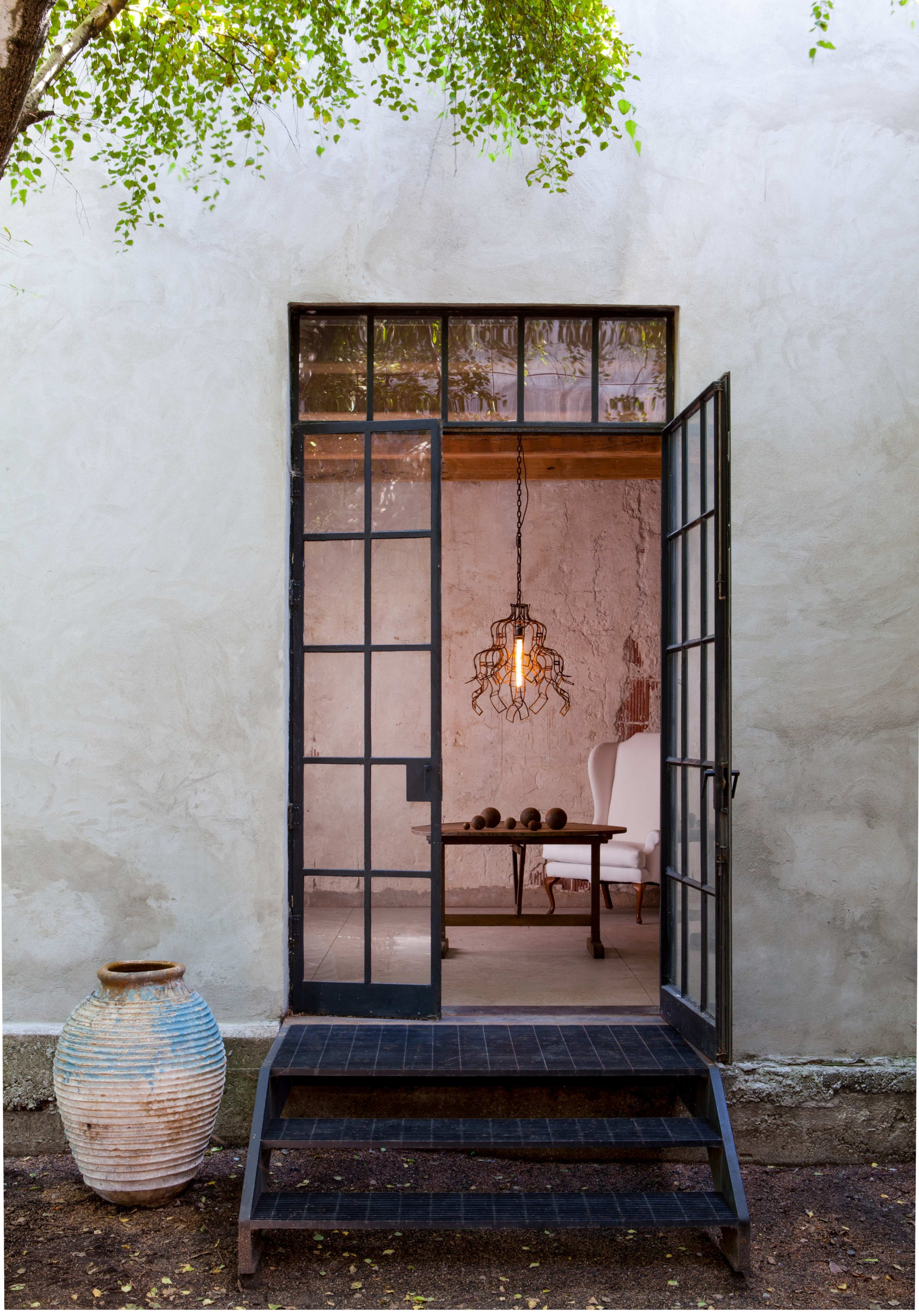 A metal-framed door opens to a minimalistic interior featuring a chair and a hanging light fixture, with an ornate vase placed nearby.