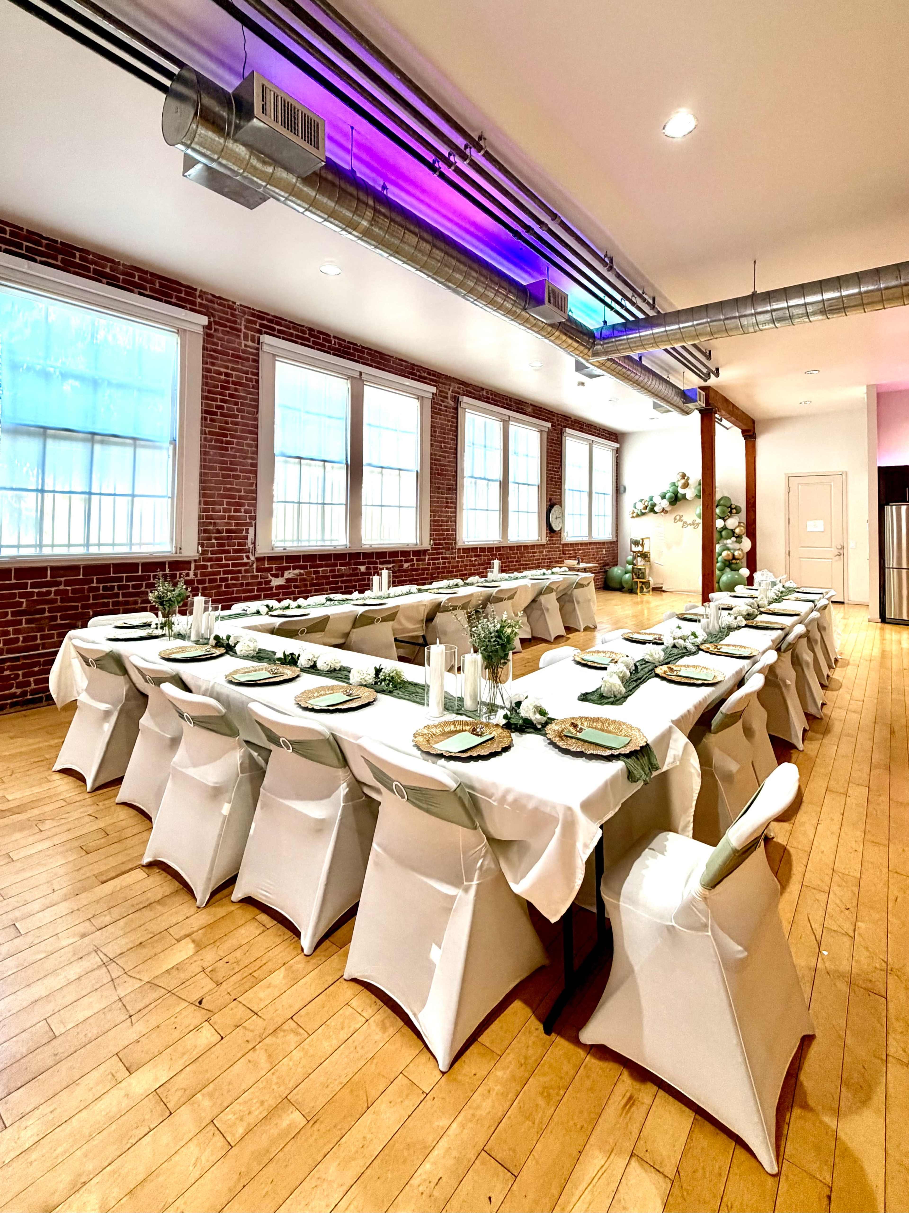 A large dining space features multiple tables arranged with white tablecloths and decorative place settings, surrounded by exposed brick walls and modern lighting.