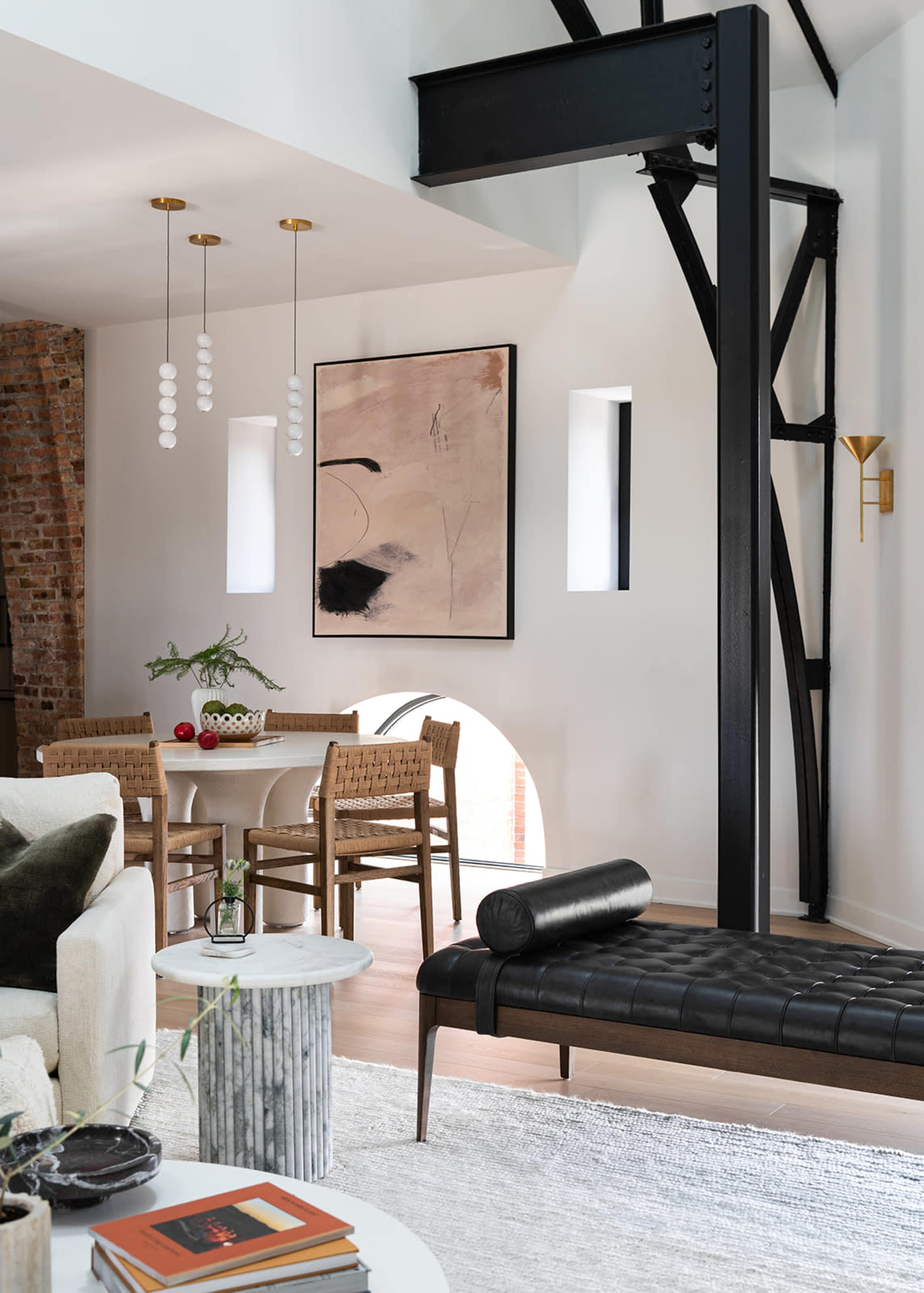 The image shows a modern living space featuring a beige sofa, a black bench, a dining area with woven chairs, and artistic wall decor, all complemented by industrial-style elements and large windows.
