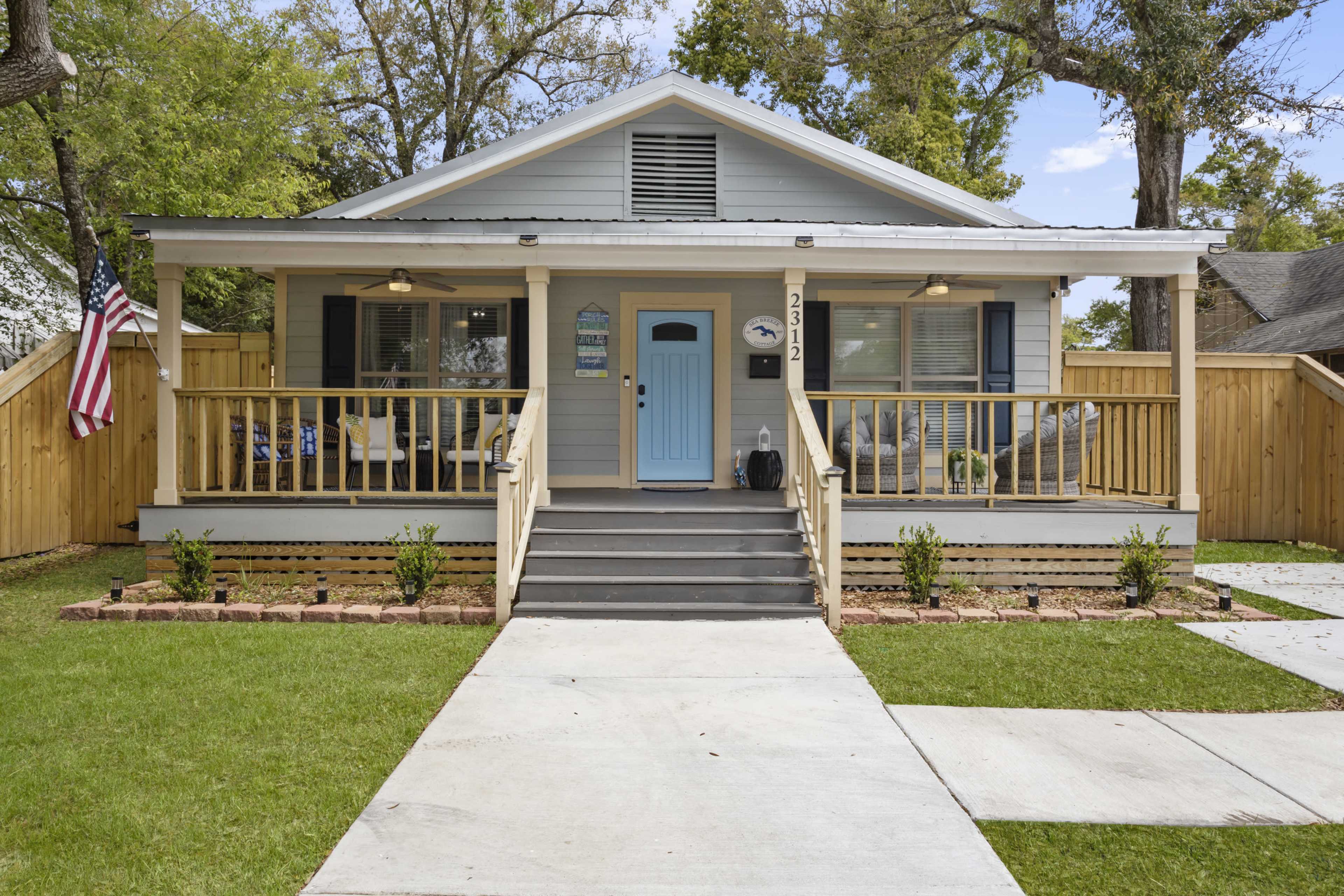 A single-story blue house with a front porch, flanked by small shrubs and trimmed grass, featuring a light blue door and an American flag.
