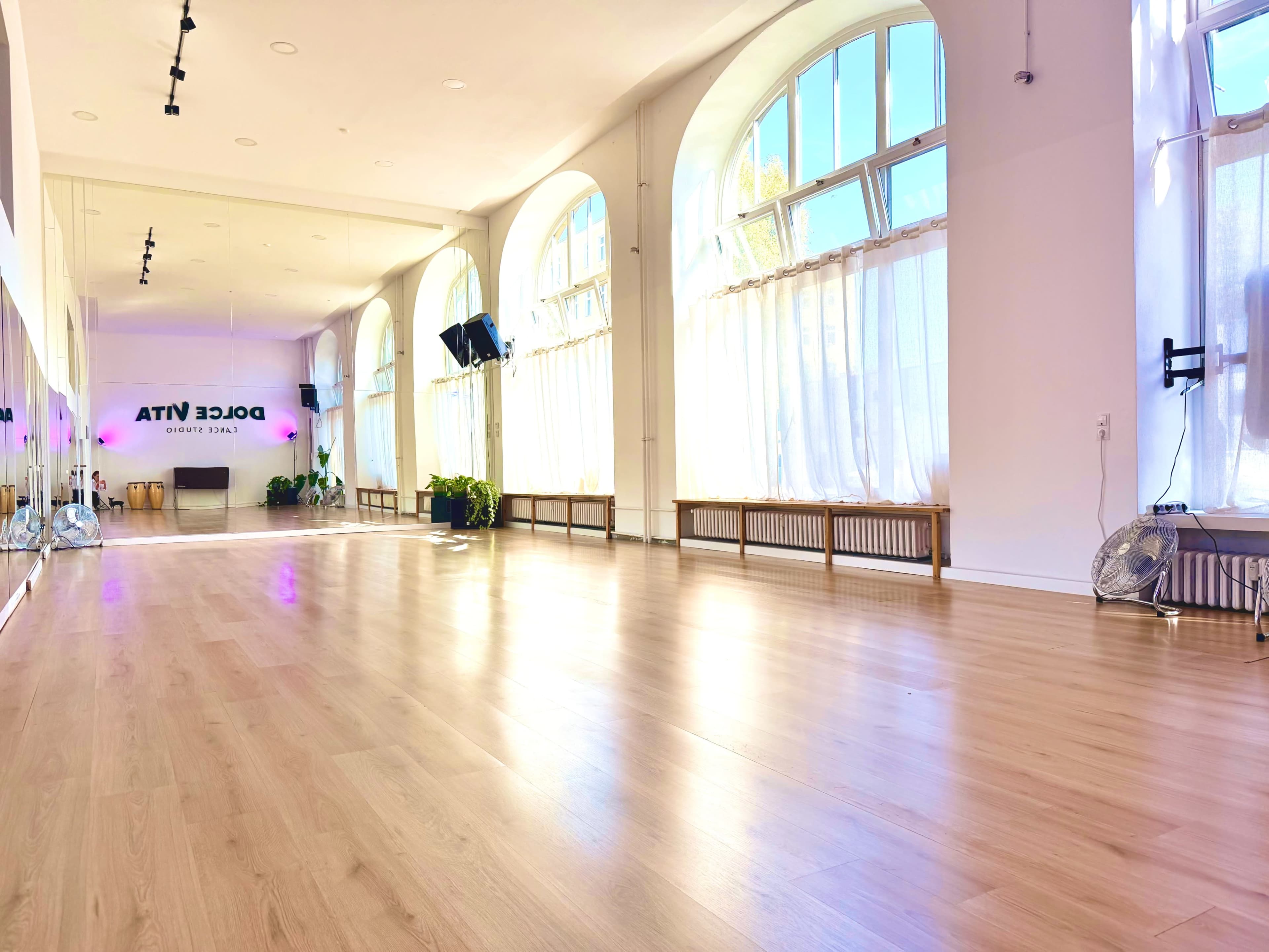 The image shows a spacious dance studio with large windows letting in natural light, featuring wooden flooring and minimal decor.