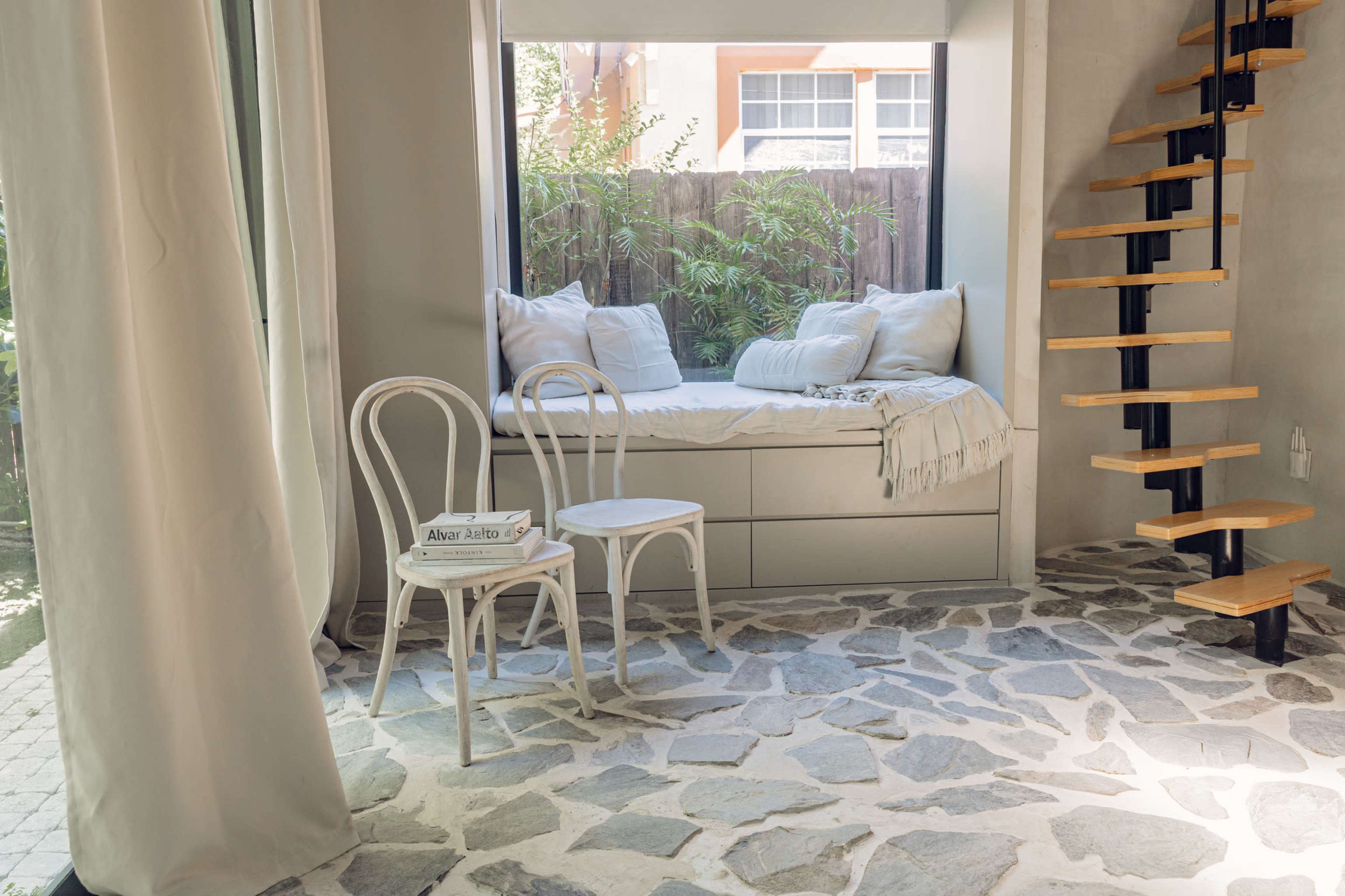 The image shows a cozy nook with a window seat adorned with cushions, accompanied by two white chairs and a spiral staircase nearby, all set against a stone floor.